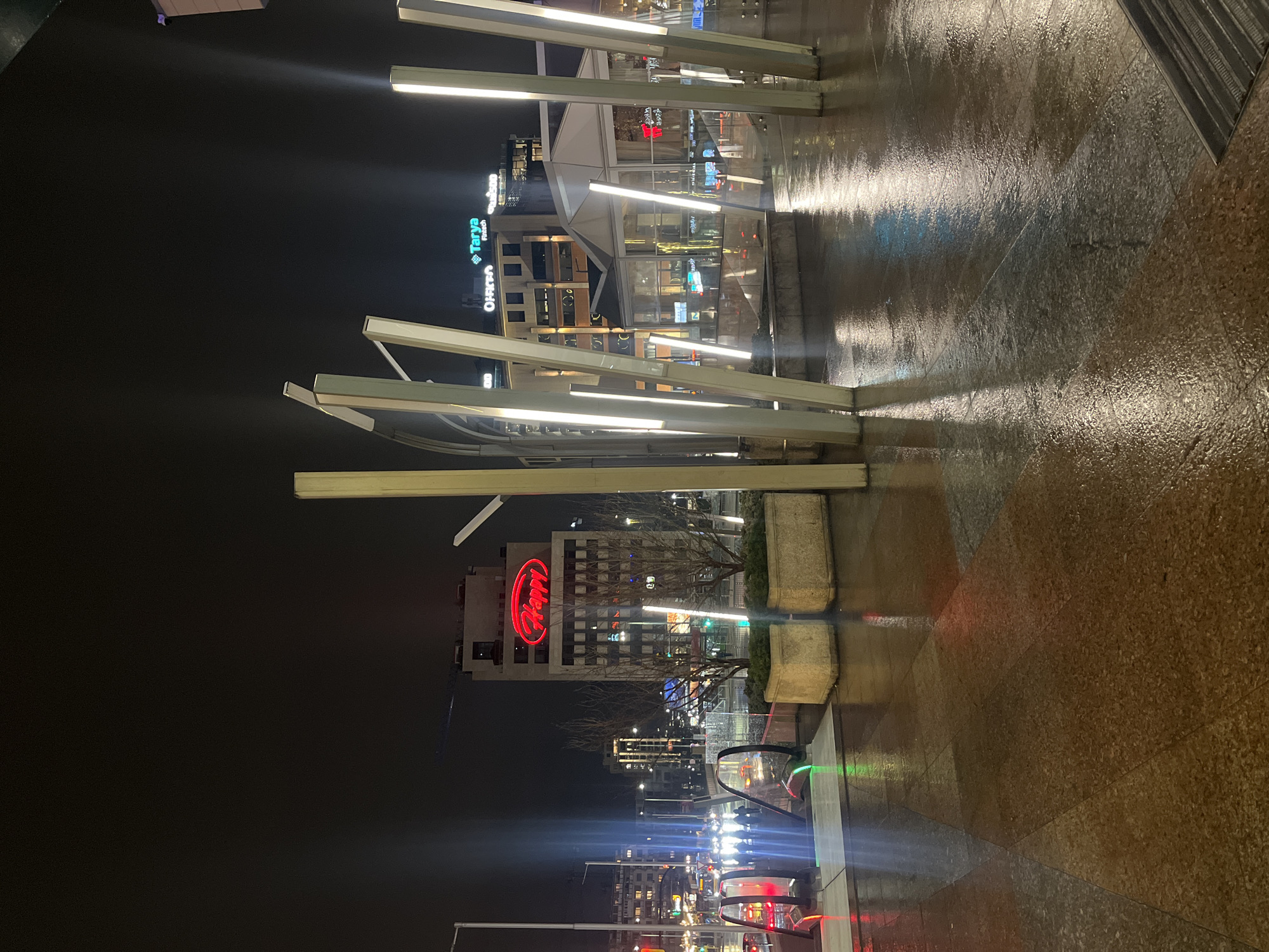 The image shows an urban scene at night in Sofia, Bulgaria. The foreground features a wet, patterned pavement reflecting light. Several modern, angled light installations stand on the pavement. In the background, there are buildings with various illuminated signs, including "Tanya" and a large, red "Happy" logo. Streetlights and vehicle lights indicate active streets. The environment suggests a business or commercial district. There are no people visible.
