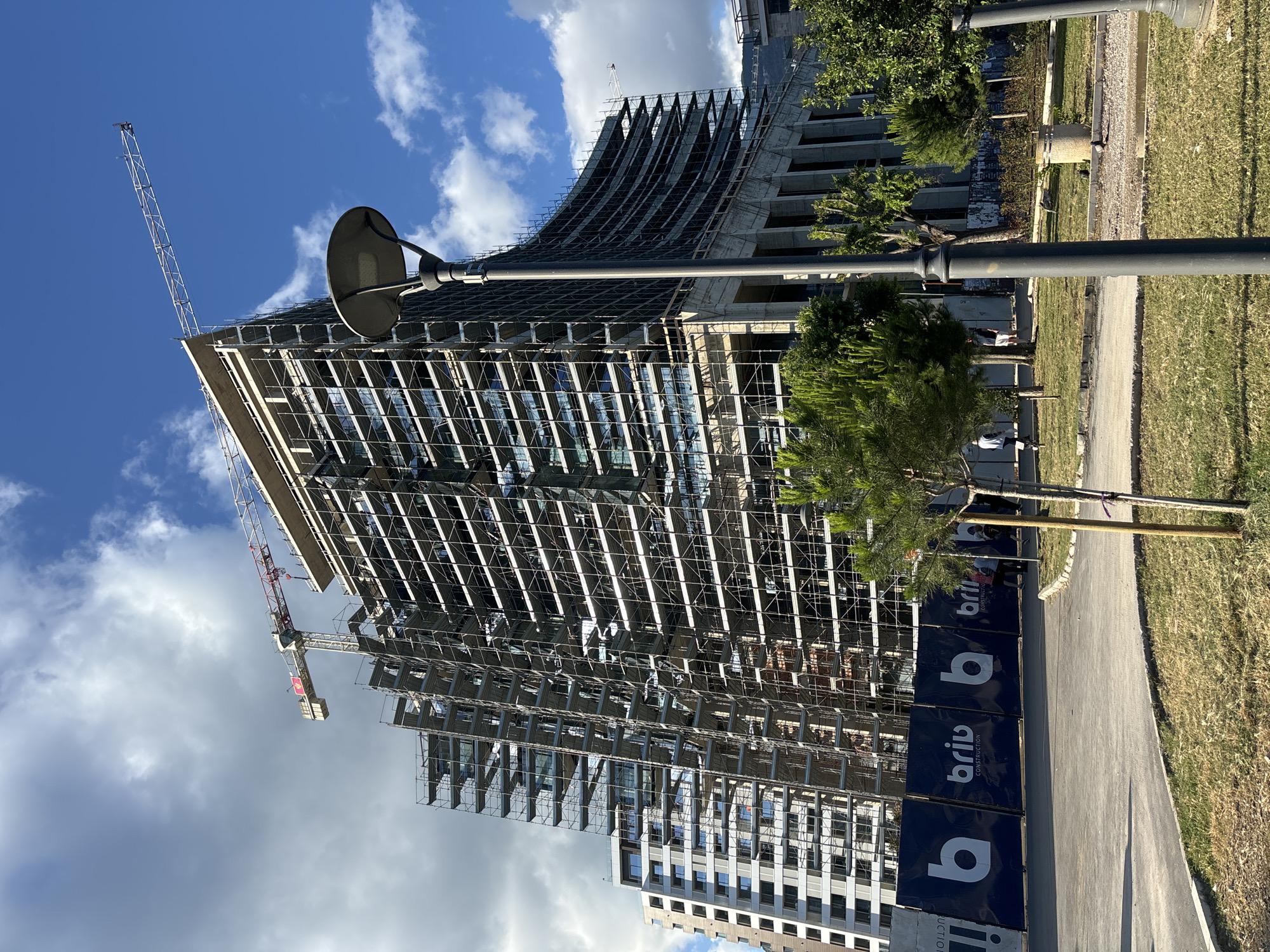The image displays a tall, modern skyscraper under construction, set against a bright blue sky dotted with fluffy white clouds. The building dominates the left and central parts of the frame, extensively covered in metal scaffolding that runs across its facade. A distinctive architectural element is visible near the top of the structure, featuring a curved, wave-like concrete design. Below this, numerous glass panels or reflective surfaces are visible between the scaffolding, indicating a contemporary design. A tall red and white tower crane is positioned to the left of the building, its long arm extending upwards into the sky. In the lower right background, another multi-story building, appearing more complete with multiple windows, can be seen.
In the foreground and extending along the right side of the image, a paved path or sidewalk runs parallel to the construction site, bordered by a strip of green grass. Several trees with dark green foliage are scattered along this grassy area. A large, dark streetlamp with a circular, dish-like head stands prominently in the mid-ground, partially obscuring the building.
Along the perimeter of the construction site, a series of dark blue hoarding panels are visible. These panels feature white text that clearly reads "brib CONSTRUCTION," alongside a stylized white 'b' logo. The scene is illuminated by bright daylight, suggesting a sunny day. No individuals are prominently featured, but the extensive scaffolding and the tower crane clearly indicate ongoing construction activity.