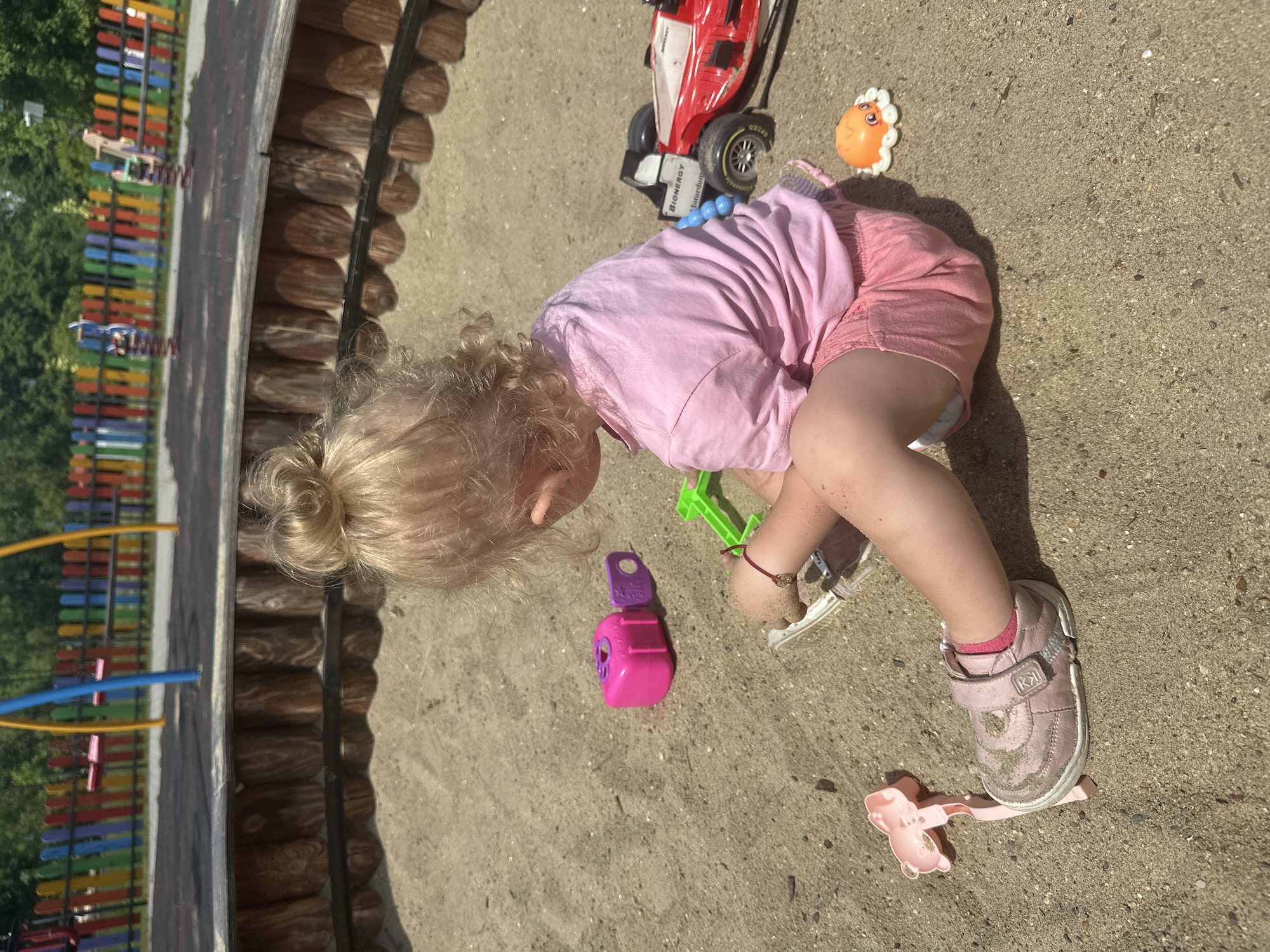 A young child with blonde hair pulled into a bun is sitting in a sandbox, facing away from the camera. The child is wearing a pink t-shirt and pink shorts, and pink shoes. The child's left hand is in the sand, and they appear to be playing with sand. Several toys are scattered in the sandbox around the child, including a pink toy, a green shovel, a red and black toy car, and a small orange octopus toy. In the background, a wooden border surrounds the sandbox, and beyond that, a colorful fence and playground equipment are visible, suggesting the location is a public playground. The overall setting is outdoors during the daytime, with bright sunlight casting shadows. The scene is consistent with a playground in Donchovci, Bulgaria, given the typical features of such facilities.