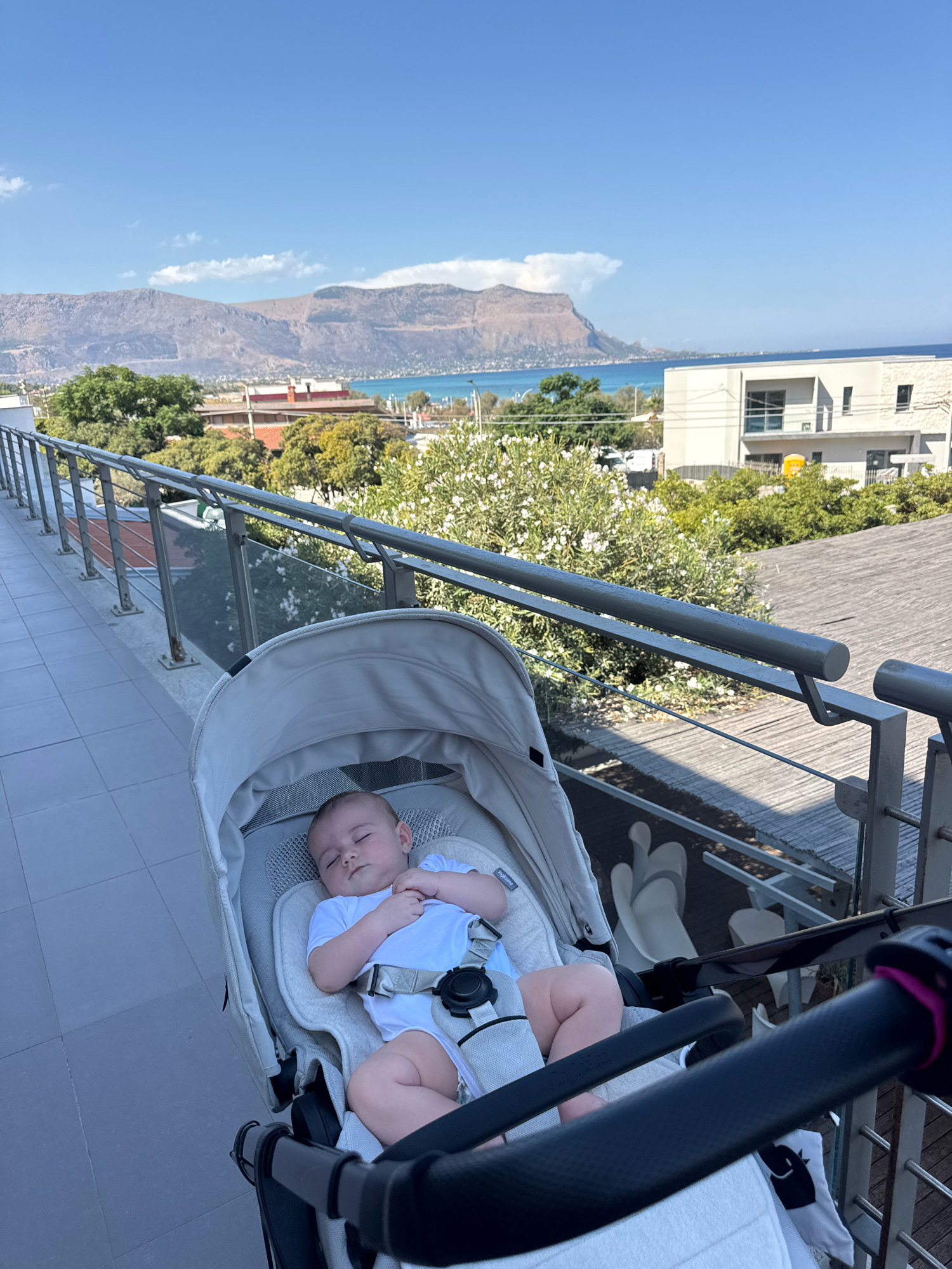 A baby is sleeping in a stroller on a balcony overlooking a coastal city. The baby is wearing a white onesie and is secured by a harness. The stroller is light gray. In the background, there are buildings, trees, and mountains. The sky is blue with a few white clouds. The scene appears to be in Capaci, Italy.