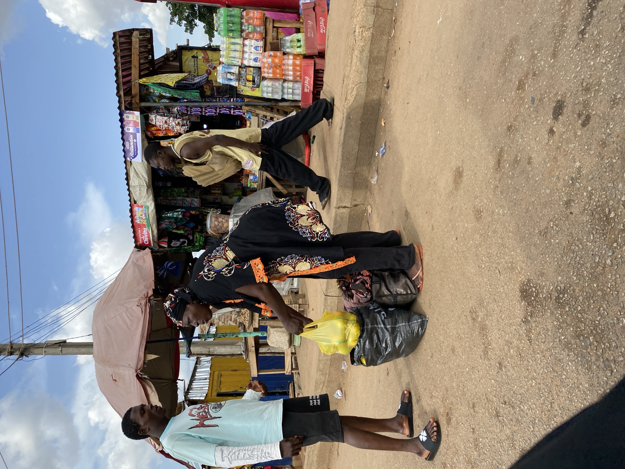 A street scene in Bukuru, Nigeria, presents three individuals at a roadside stall. The stall displays a range of goods, including beverages and snacks. A man in a yellow shirt stands in front of the stall. A woman in a black patterned dress bends over to adjust a yellow bag on the ground, where a black plastic bag rests. A younger man in a light blue shirt and dark shorts stands nearby. The ground is paved and shows signs of wear. Overhead, an umbrella provides shade. The sky is partly cloudy.