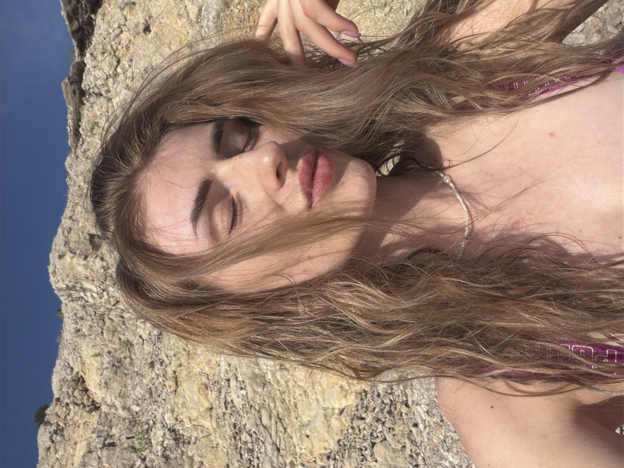 In a selfie taken outdoors, a young woman with long, wavy brown hair is captured with her eyes closed and lips puckered. She is wearing a purple swimsuit with a beaded necklace. The background consists of a rocky cliff face and a clear blue sky. The lighting suggests it is daytime, likely sunny. The woman's hand is raised, gently touching her hair. The overall impression is one of relaxation and enjoying the outdoors.