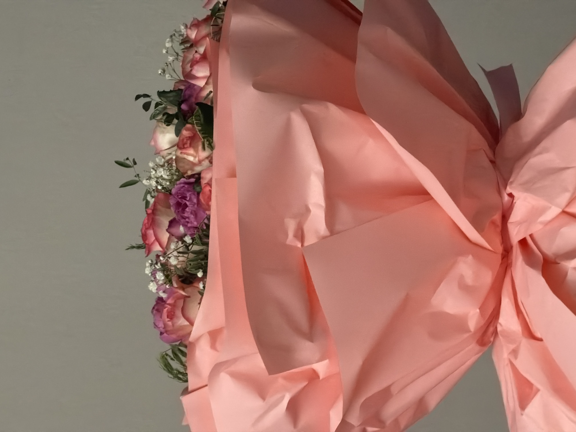 The image is a close-up shot of a bouquet of flowers, predominantly pink and white roses, with some purple accents and small white filler flowers. The bouquet is wrapped in crinkled, light pink paper, creating a soft, voluminous presentation. The background is a plain, light grey wall. There are no people or specific landmarks visible to indicate a location in Yerevan, Armenia, or any other city. The focus is entirely on the floral arrangement and its packaging.