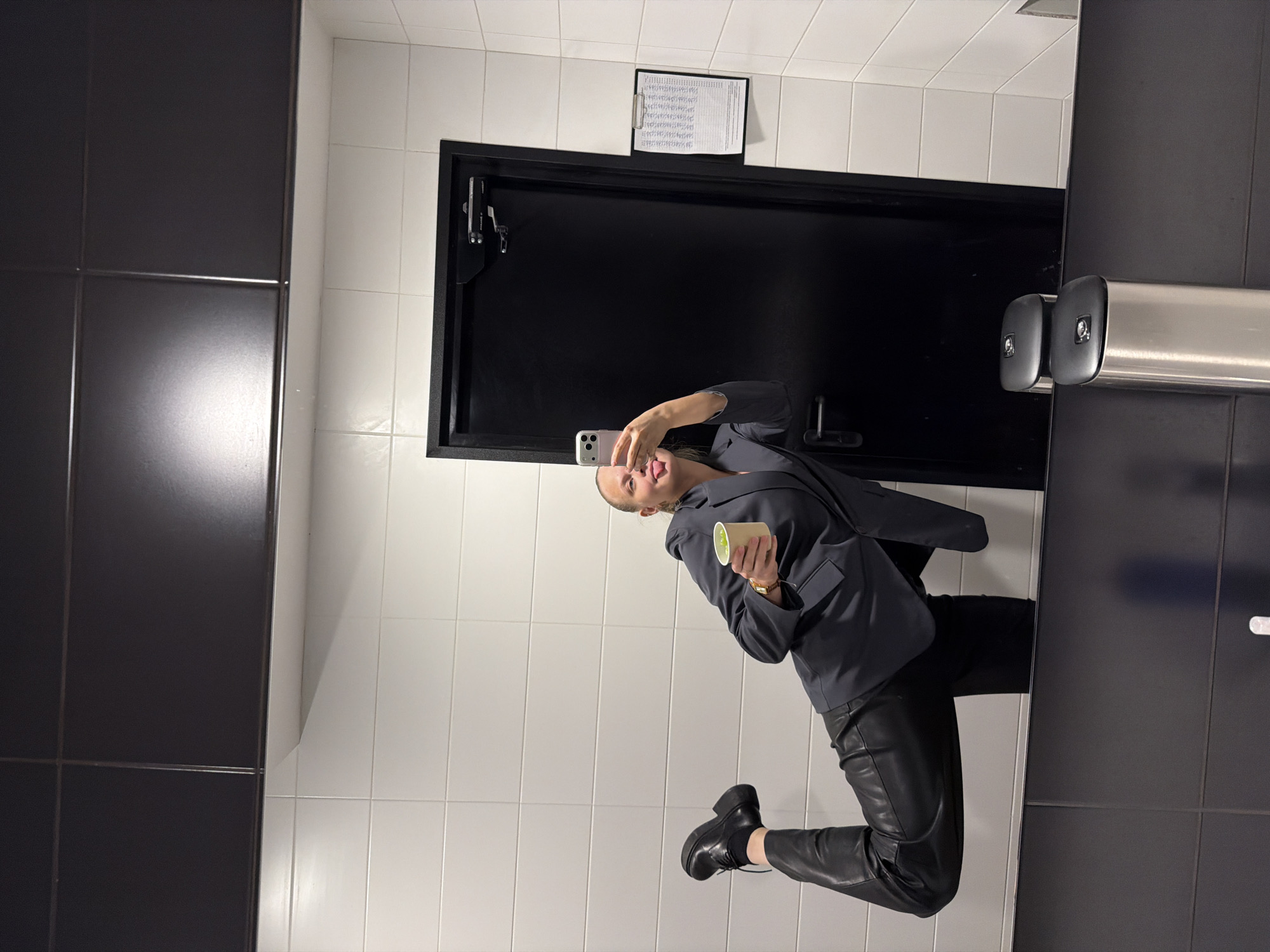 A person is visible, taking a selfie in a bathroom. The person is wearing a dark grey blazer, black leather pants, and black boots. They are holding a cup in their left hand and a phone in their right hand, sticking their tongue out. The bathroom has white tiled walls with black tiled accents. A black door is behind the person, with a clipboard attached above it. A metal toilet paper dispenser is visible to the right. The exact location in Izhevsk, Russia cannot be determined from the image.