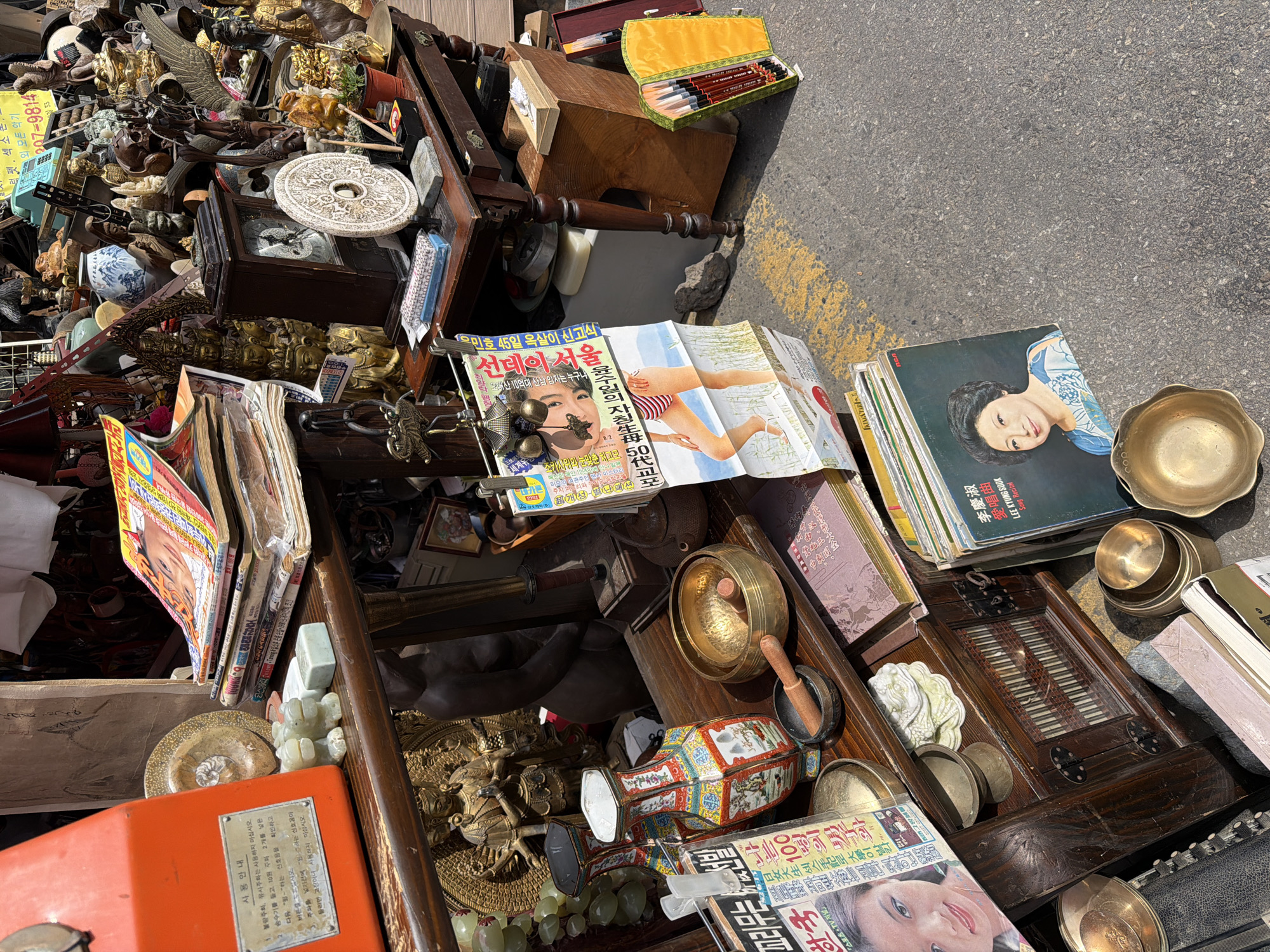 The image captures a collection of items for sale at a street market in Seoul, South Korea. The goods, including books, records, magazines, and various brass and decorative objects, are piled on makeshift displays, seemingly on the street. The magazines and records feature Korean text and images, indicating their origin and target audience. The overall impression is of a bustling open-air market with a wide array of vintage and potentially antique items.
