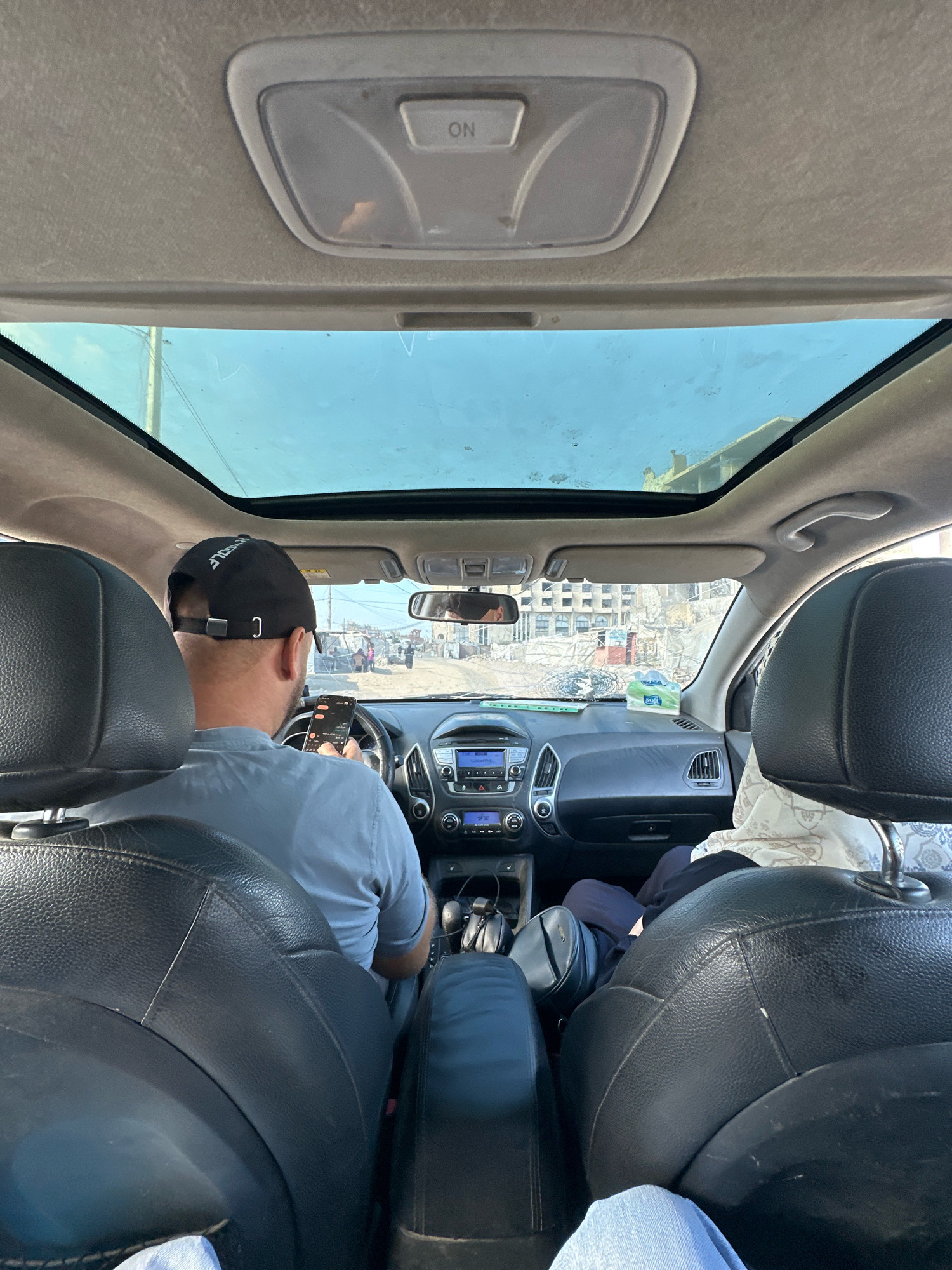 An interior shot of a car driving through the city of Gaza. The camera is positioned looking forward from the back seat, showing the driver and a passenger in the front. The driver, wearing a black baseball cap and a light blue t-shirt, is holding a smartphone. The passenger, wearing a headscarf, is seated to the right of the driver. A large sunroof is open, revealing a clear sky. The car's dashboard is visible, with a radio and air conditioning controls. Outside the car, the cityscape of Gaza is visible, with buildings and a street with people and vehicles. The overall atmosphere is one of everyday life in a busy urban environment.