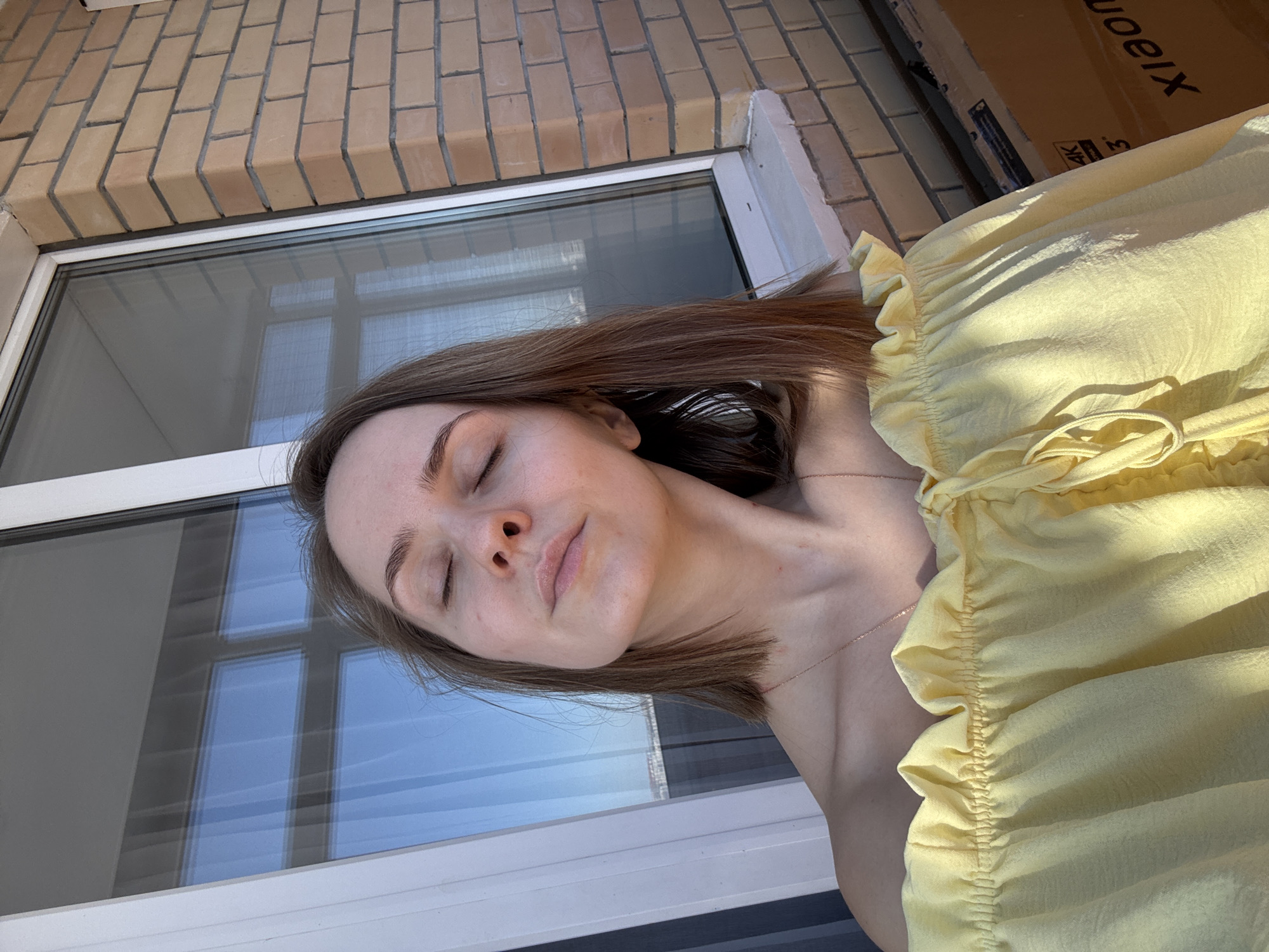 A female subject with shoulder-length brown hair is positioned in the foreground, eyes closed, face tilted slightly upward and to the right, bathed in direct sunlight. She wears a light yellow, off-the-shoulder ruffled top, revealing a thin golden-colored chain necklace on her bare clavicle area. The background features a large window with white framing on the left, reflecting a clear blue sky and a distant urban landscape. To the right of the window, a section of a beige brick wall with light grey mortar is visible. A white window sill or ledge is present below the window. In the lower right corner, a brown cardboard box is partially visible, displaying the text "4K" and the partial word "xiaomi". The scene is situated in the city of Perm, Russia, likely within a residential interior or on a balcony/loggia.