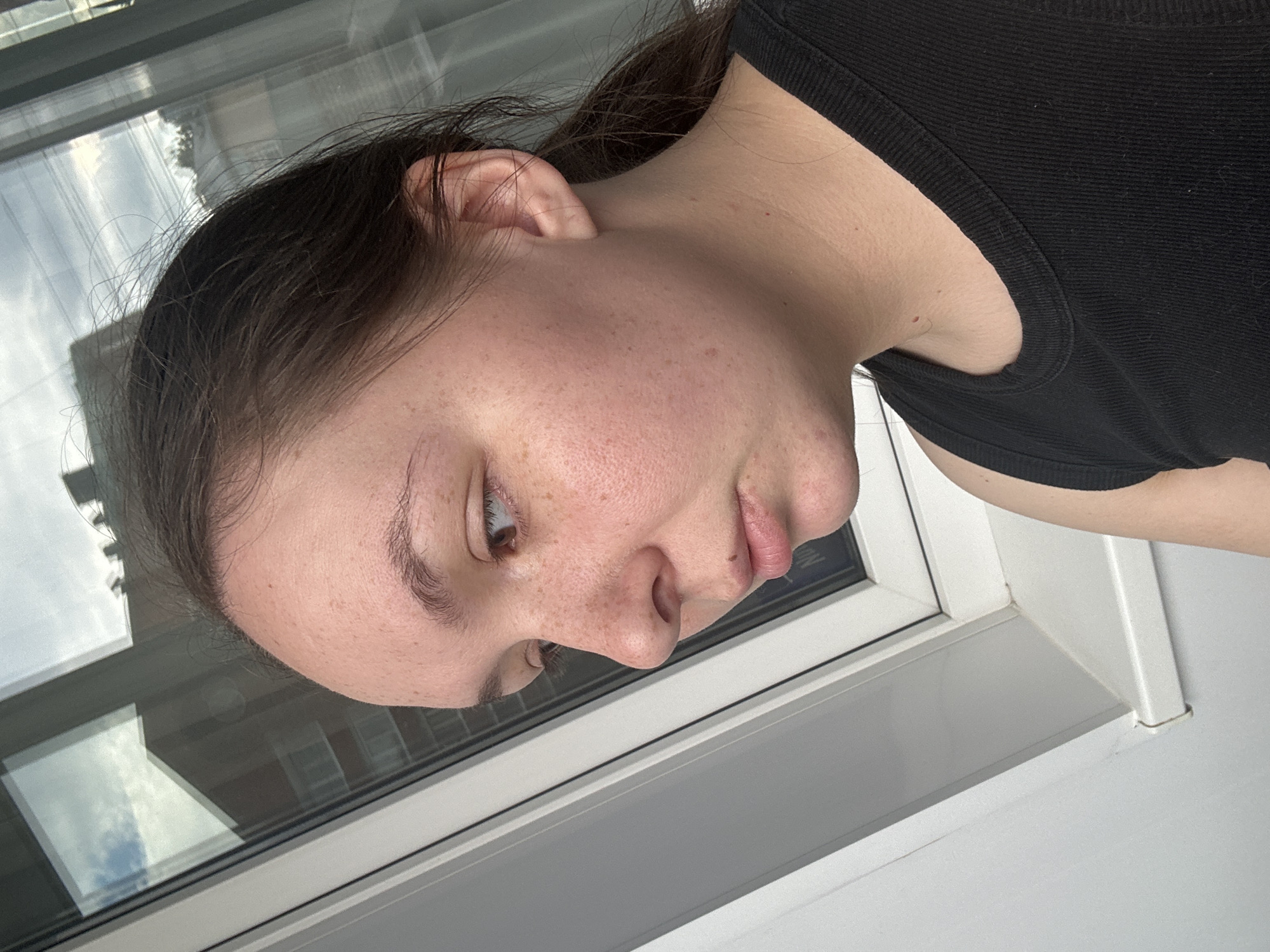A young woman with brown hair pulled back in a ponytail and freckled skin is shown in a close-up profile shot. She is wearing a black tank top and is looking out of a window. The window is large and reflects buildings and the sky, suggesting an urban setting. The woman's facial expression is neutral, and she is positioned with her head tilted slightly downwards. The lighting appears natural, coming from the window. The background inside the room is not clearly visible, but the focus is on the woman and the window. The overall scene suggests a moment of quiet observation or reflection. This image was captured in Chaikovsky, Russia.