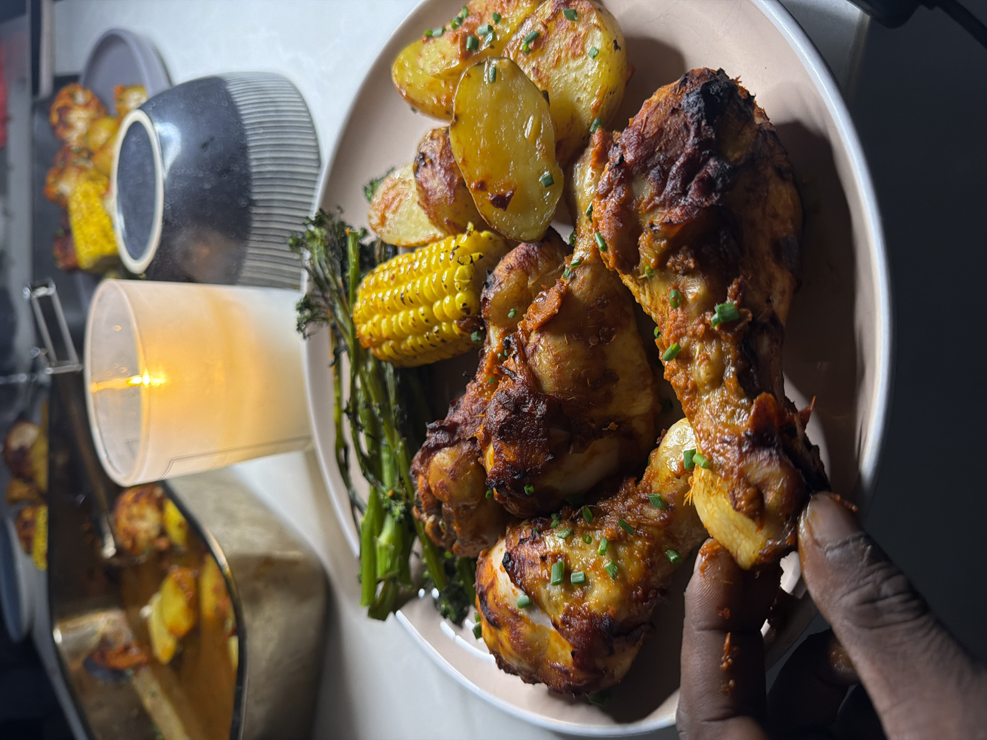 An overhead, close-up shot shows a person's hand reaching for a chicken drumstick from a plate of food. The plate contains several chicken drumsticks, roasted potatoes, and grilled broccoli, all garnished with chopped chives. In the background, a lit candle sits beside a dark, ribbed bowl, and a roasting pan filled with more food is visible. The scene appears to be set on a countertop or table, with a blurred background suggesting an indoor setting.