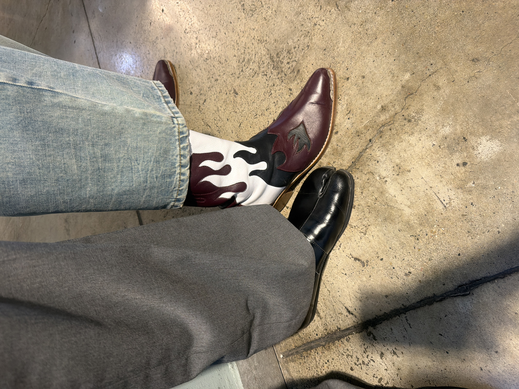 The image captures a close-up view of two individuals' legs and feet, showcasing their distinct footwear. On the left, a person wears dark grey wide-leg trousers and black split-toe shoes, a style often referred to as tabi shoes. The shoe is a flat, slip-on design with a glossy finish. To the right, another individual is dressed in light-wash denim jeans and a pair of striking burgundy and white cowboy boots adorned with a flame design in black and dark red. The boots have pointed toes and a visible wooden heel. Both individuals are standing on a textured concrete surface, which appears to be a sidewalk or ground-level area, possibly in an urban setting. The lighting suggests an outdoor environment, with shadows cast on the ground. While the specific location in Tbilisi, Georgia, cannot be definitively identified from the image alone, the urban setting and concrete ground are consistent with many public spaces within the city.