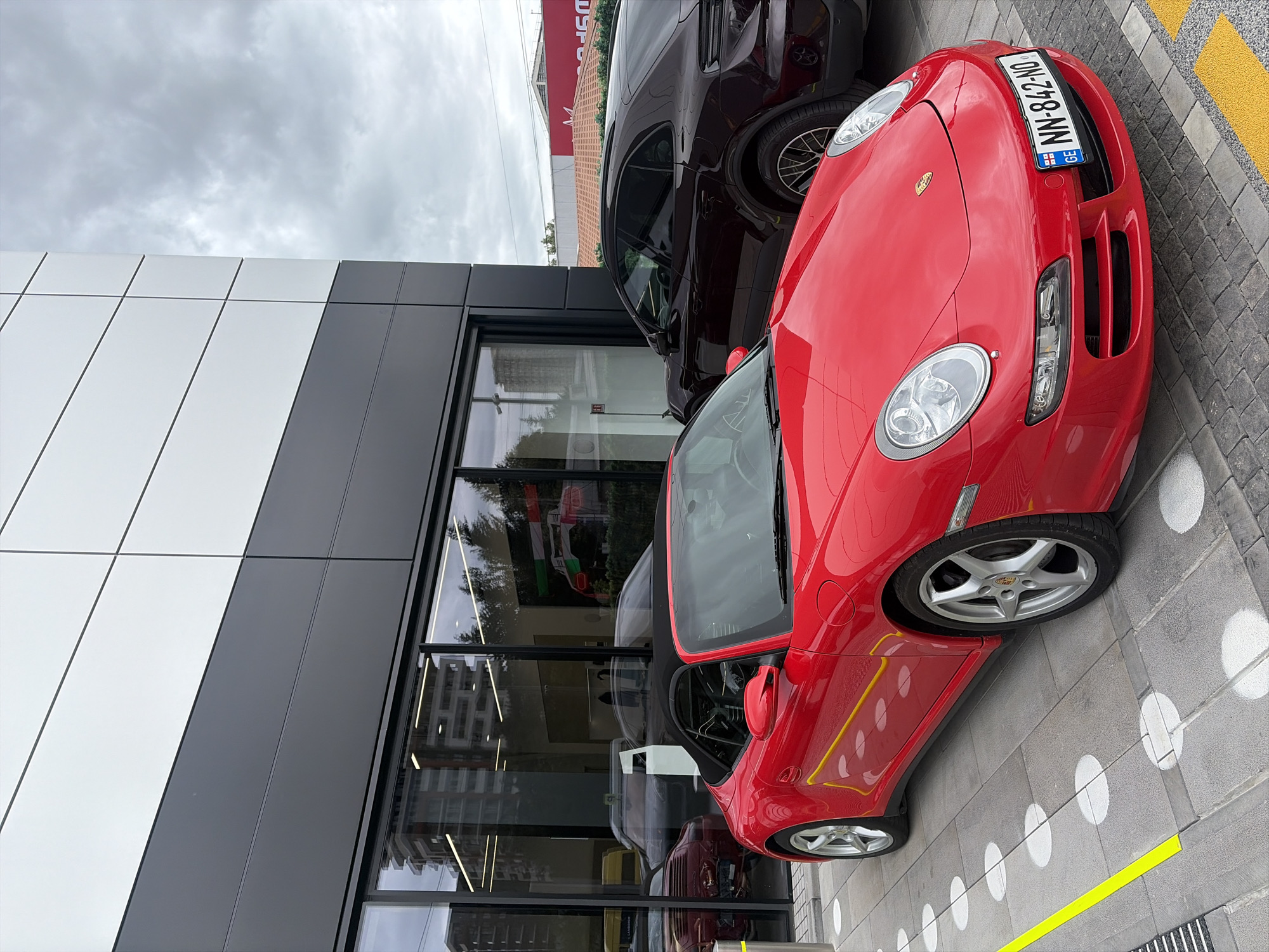 The image depicts a red Porsche parked outside a modern building with gray and white panels and large windows. Next to it is a dark-colored SUV. The location is identified as Tbilisi, Georgia, based on the license plate of the red Porsche. The pavement is decorated with white circles and yellow lines. The sky is overcast. The reflection in the building's windows suggests the presence of other vehicles inside.