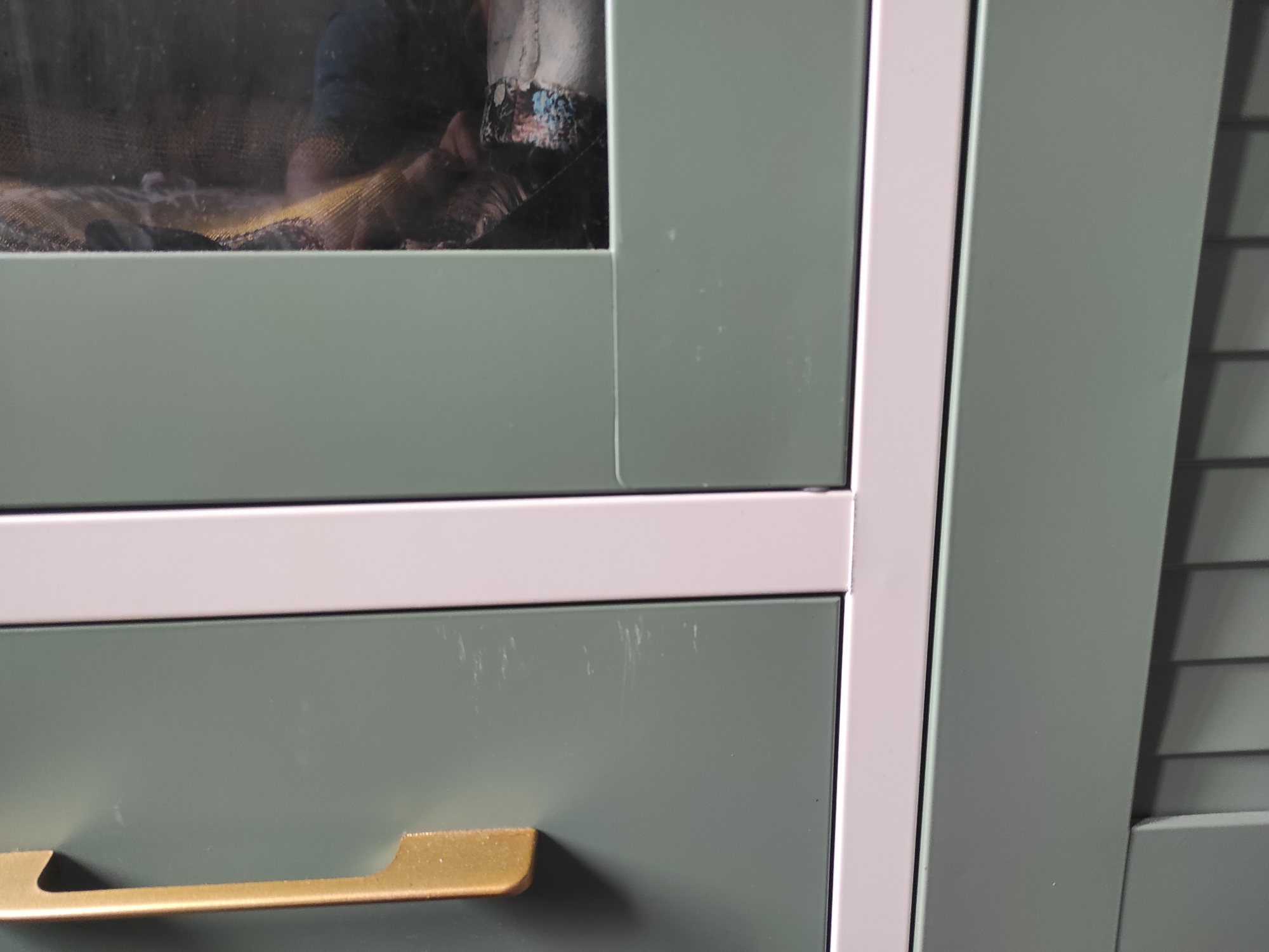 The image shows a close-up of a pale green cabinet with a white trim. The cabinet has a glass-fronted upper section, partially obscured by reflections, showing what appears to be a person wearing a dark shirt and some patterned fabric. Below the glass section is a drawer with a gold-colored handle. The right side of the image shows a section of the cabinet with horizontal slats, suggesting it might be a shutter or ventilation. The scene is indoors, with no visible elements that would place it specifically in Muara Jawa Ulu, Indonesia. The focus is on the texture and details of the cabinet's surfaces, with some minor scuff marks visible on the painted surfaces.