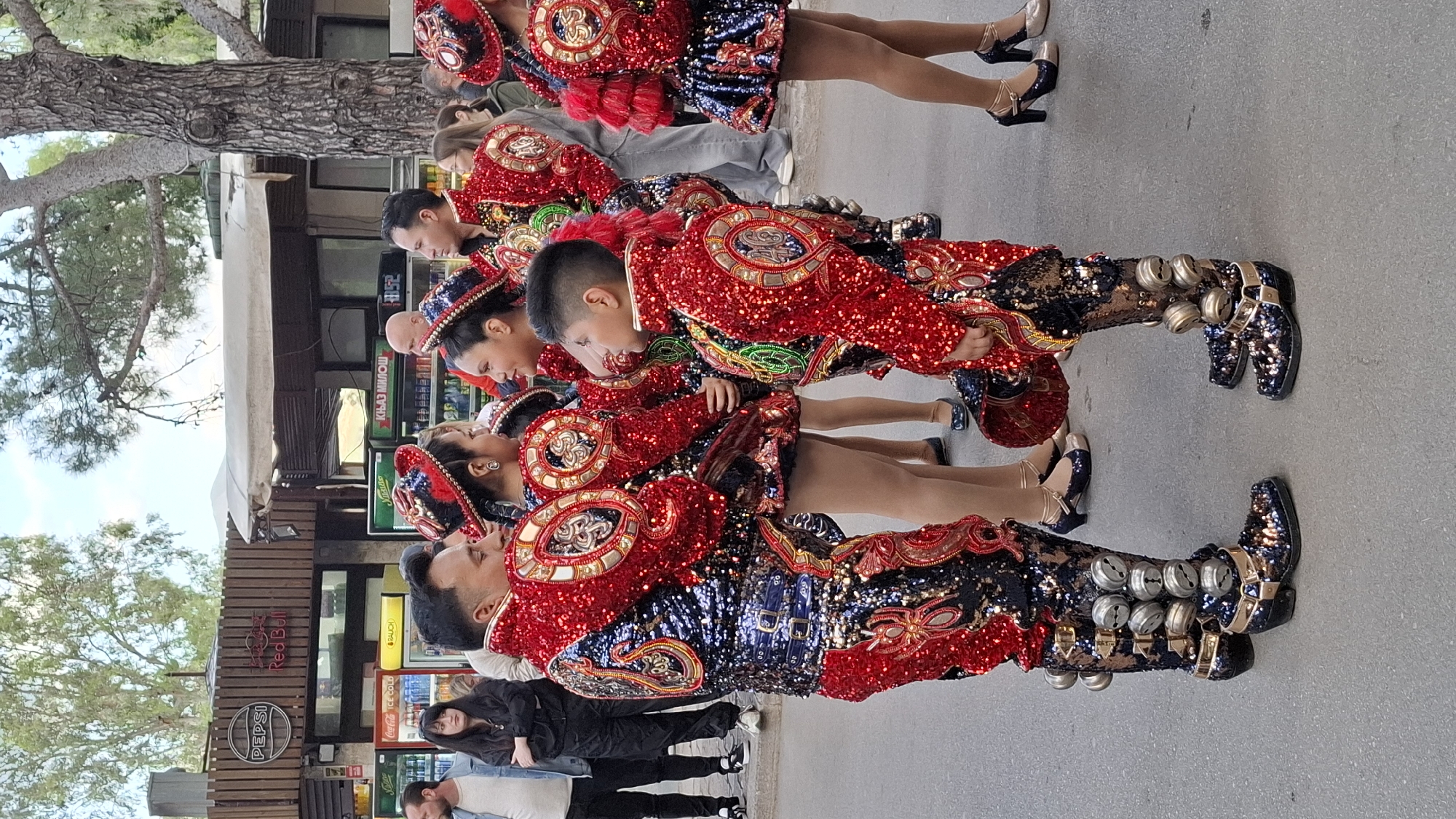 A group of people are seen in traditional costumes. The costumes are vibrant and decorated with sequins, in colors such as red, blue, and gold. The dancers are wearing hats decorated with feathers and some have bells attached to their boots. They are standing on a paved area, likely a street or a plaza. Behind the dancers, there are buildings, including a cafe with signage for Pepsi, Coca-Cola, and Red Bull. People in casual clothing are visible in the background. The setting appears to be a street in Budva, Montenegro.