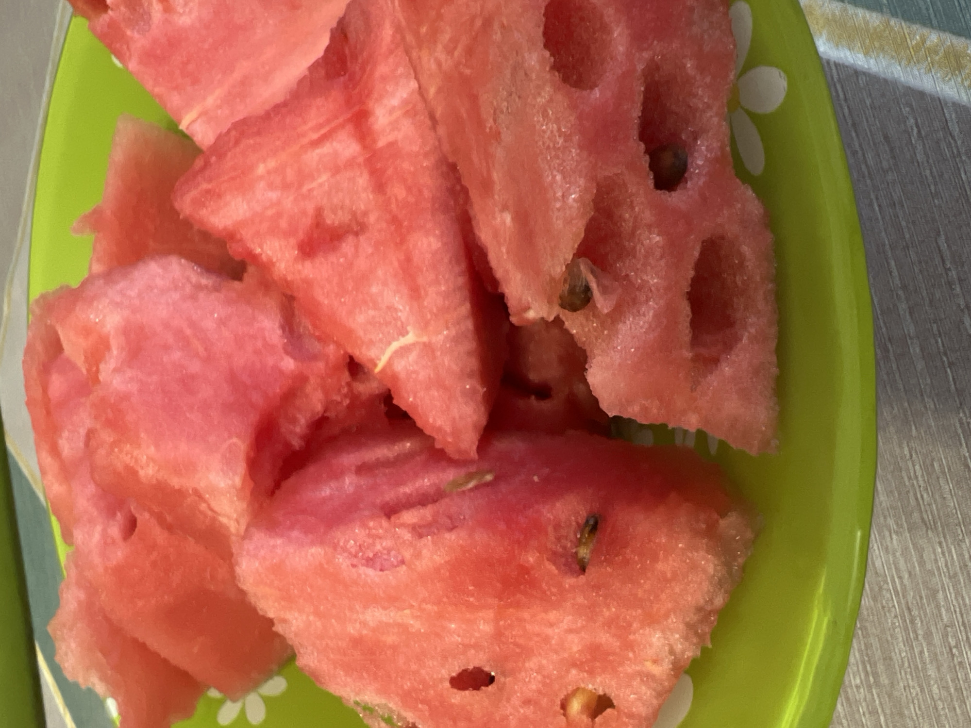 The image displays several irregular pieces of bright pinkish-red watermelon arranged on a vibrant lime green plate. The watermelon pieces vary in size and shape, showing the juicy, slightly textured flesh characteristic of the fruit. Several small, dark watermelon seeds are visible embedded within the flesh or in cavities where they might have been.
The green plate features a subtle pattern of white, stylized flowers along its rim, visible in areas not covered by the fruit. The plate rests on a surface that appears to be light-colored wood or a wood-grain patterned tabletop/mat, visible on the right side of the frame. The lighting is bright and even, suggesting an indoor setting. There are no people visible, and no discernible activity is taking place beyond the presentation of the cut fruit. No text is visible in the image. The location is Chaikovsky, Russia, though this is not visually identifiable from the image itself.