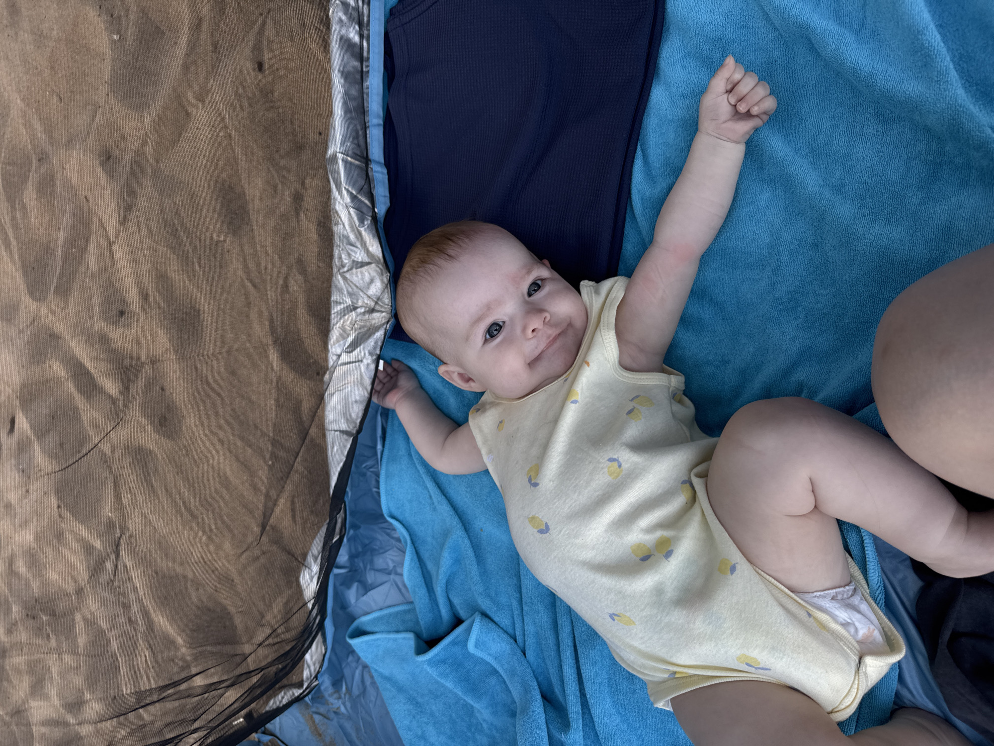 A baby lies on a blue towel, with a dark blue piece of clothing and a brown fabric with a black net beside it. The baby is wearing a yellow onesie with a lemon pattern and has its left arm raised. The scene appears to be outdoors, possibly in a park or beach area, given the brown fabric and net, suggesting a picnic or camping setting. The baby has a happy expression, looking towards the camera. The lighting suggests it is daytime.