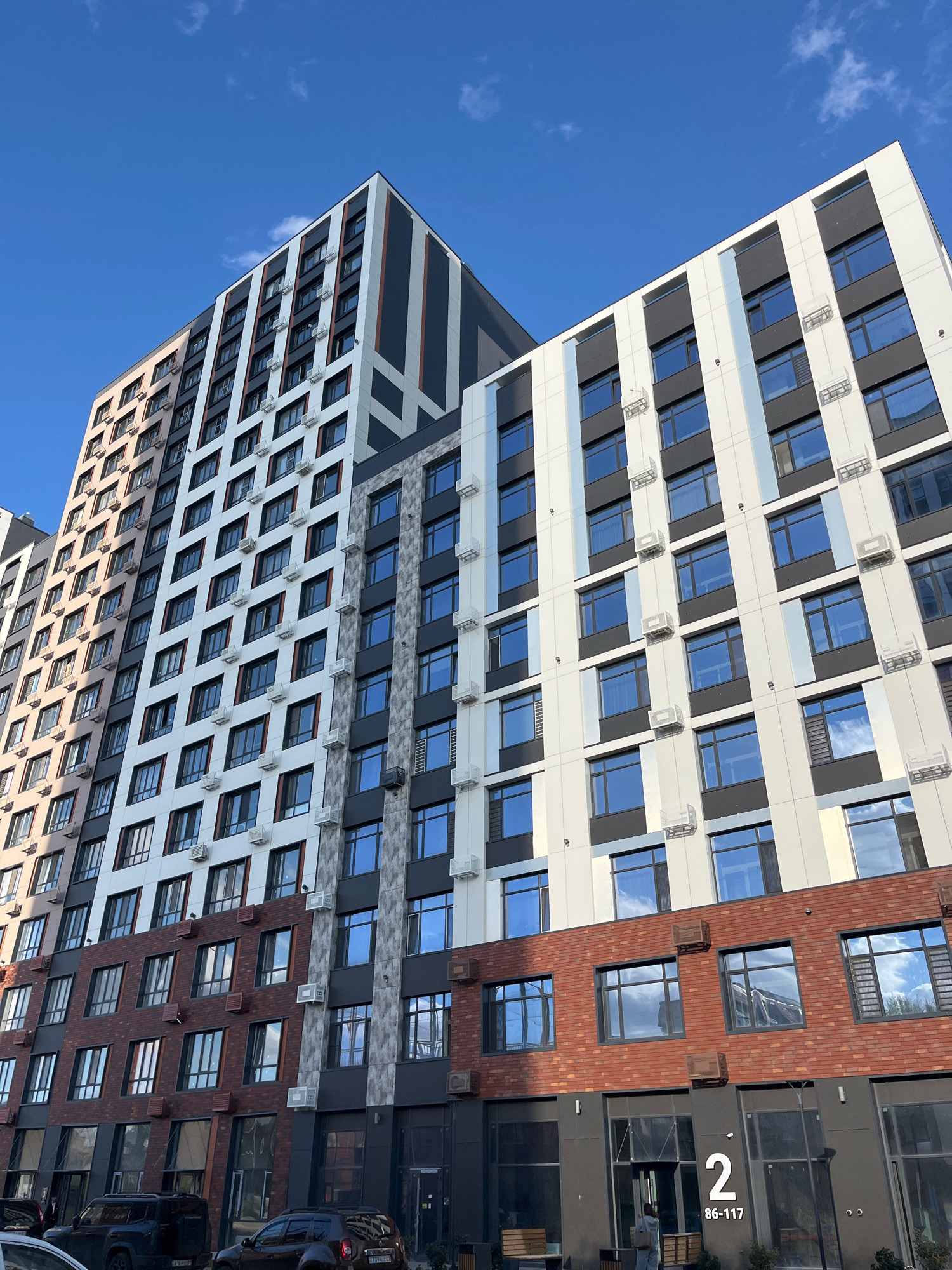 This is a low-angle, full shot of a modern, multi-story residential building complex in Astana, Kazakhstan, under a clear blue sky with scattered white clouds. The building features a contemporary design with a facade composed of white panels, dark gray accents, and red brickwork on the lower levels. Rows of windows are visible on each floor, many equipped with external air conditioning units. The building's architecture is characterized by vertical and diagonal lines, creating a dynamic visual effect. On the ground level, there are commercial spaces with large glass windows and dark gray doors. The number "2" is prominently displayed on the building, along with the alphanumeric code "86-117." Two black SUVs are parked in front of the building. The overall scene conveys a sense of urban development and modern living.