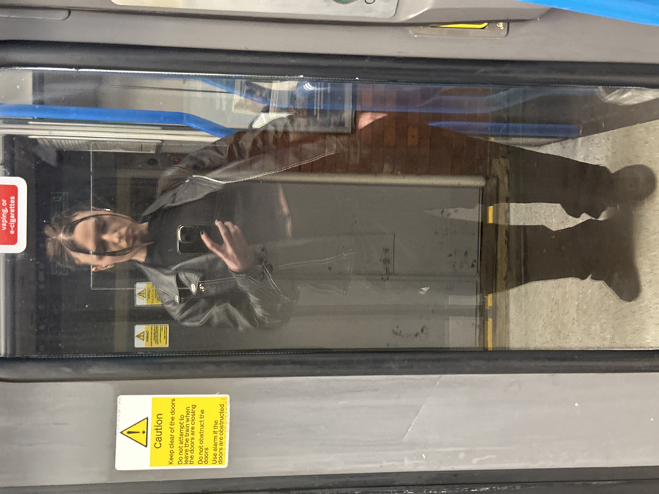 The image captures a reflection of a person in a train or bus door. The person is wearing a black jacket and trousers. They are holding a mobile phone, presumably taking a picture or video of themselves. Two caution stickers are visible on the door. One sticker advises to keep clear of the doors while they are closing and the second sticker advises against vaping and the use of e-cigarettes.