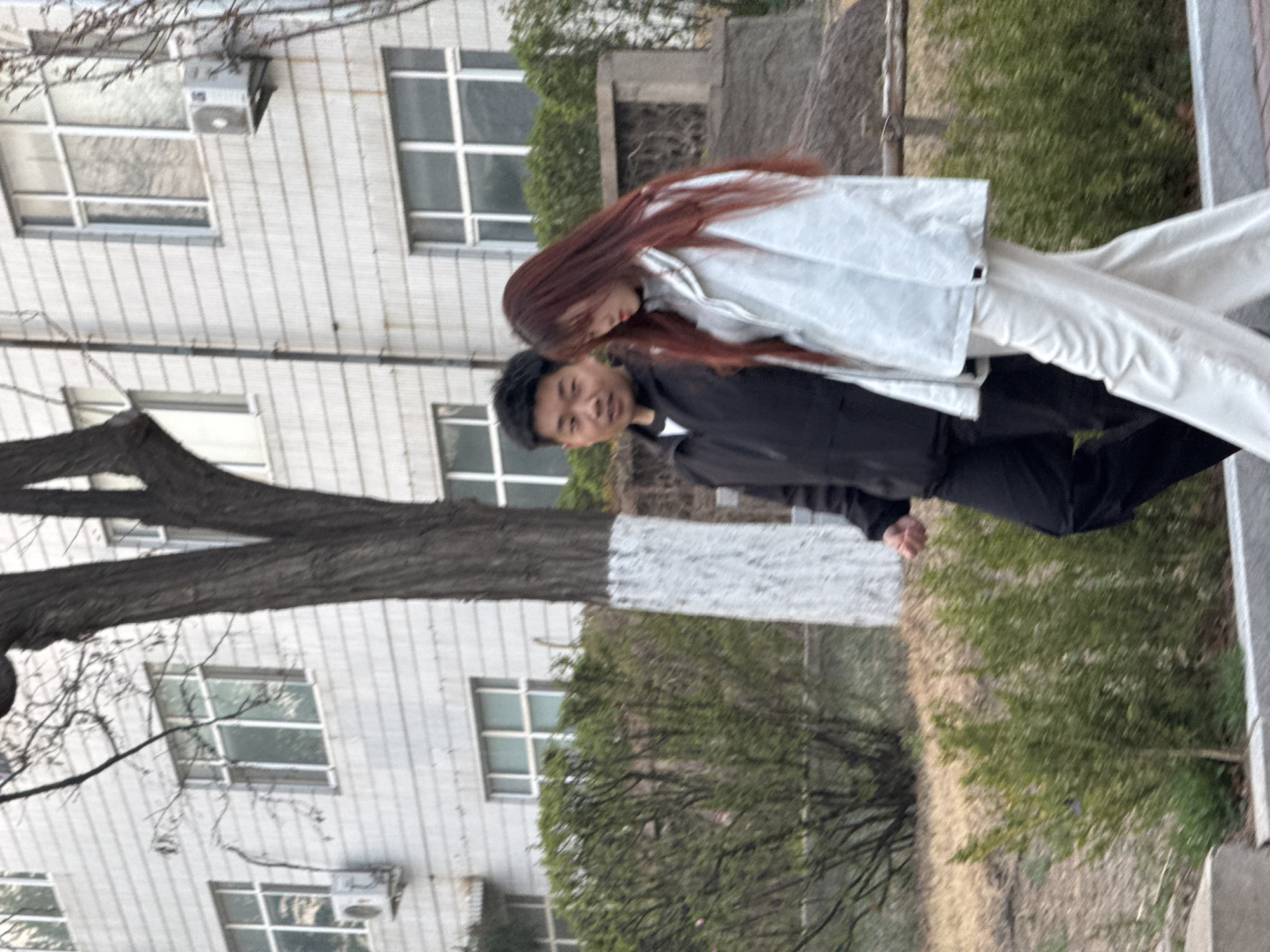 The image depicts a man and a woman standing outdoors, posing together in what appears to be Zhongtun, China. The man, positioned on the left, has short dark hair and is wearing a dark jacket and dark pants. He is looking towards the viewer with a slight smile. The woman, on the right, has long, reddish-brown hair and is dressed in a light-colored (likely white or light gray) jacket and matching pants. She is leaning her head towards the man, her face partially obscured, in a gesture suggesting affection or closeness.
They are standing in front of a light-colored building with multiple windows, some featuring multi-pane designs. An air conditioning unit is visible on the building's exterior wall. A large tree trunk, painted white around its lower section, stands in the foreground, separating the individuals from the building in some parts. Green bushes and grass are visible in the background and on the ground, along with some bare tree branches at the top of the frame. The lighting suggests it is daytime, possibly under an overcast sky or during the softer light of early morning or late afternoon. There is no visible text in the image.