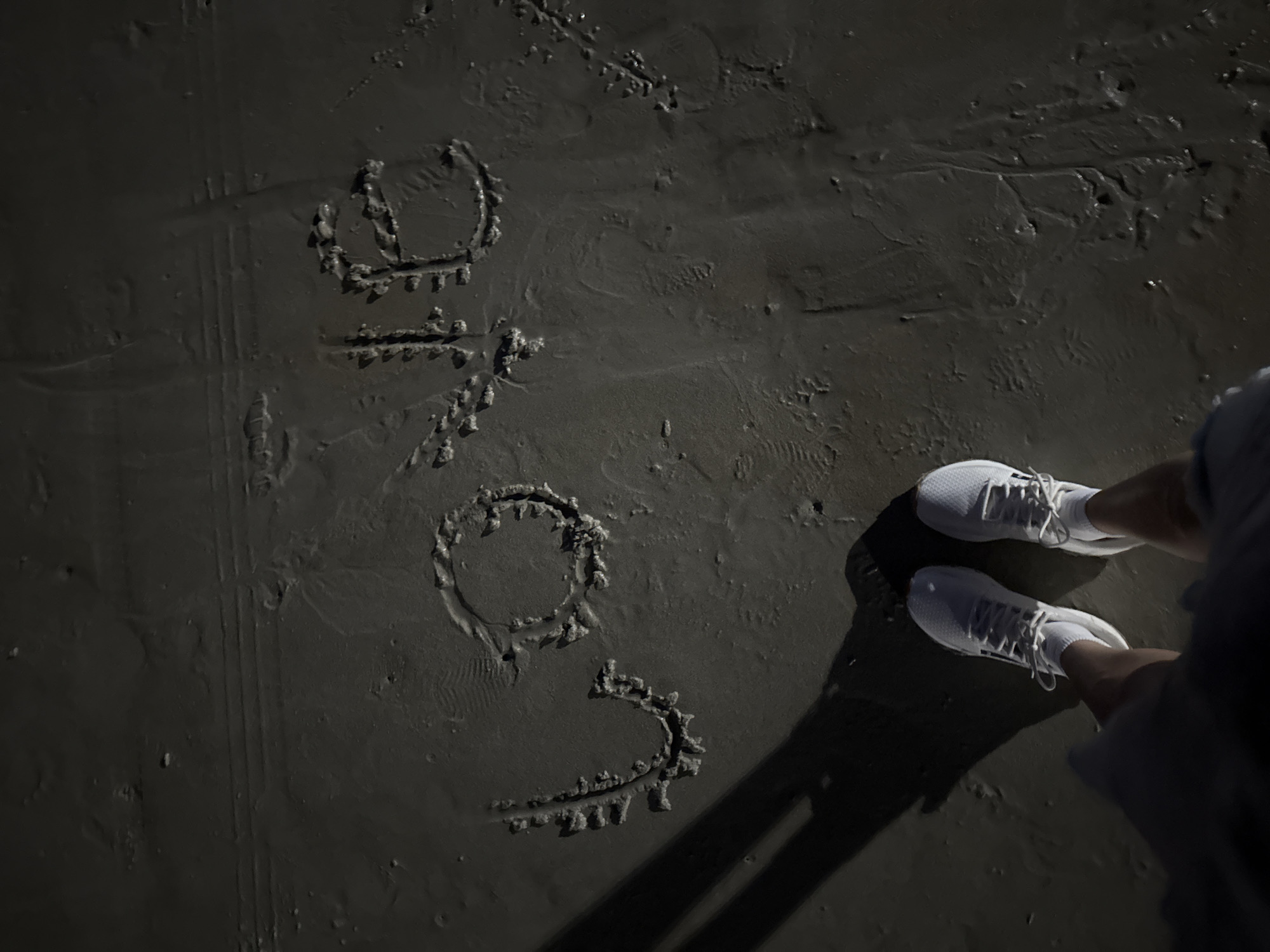 The image depicts the word "LOVE" written in the wet sand on a beach. The letters are formed with distinct impressions, suggesting they were drawn by hand. The sand appears dark and wet, with ripples and texture from the tide or footprints. In the lower part of the frame, a person's legs and feet are visible, wearing white sneakers and socks. The person is casting a long shadow to the left, indicating a low sun angle, likely during sunrise or sunset. The setting appears to be a beach, consistent with Karon, Thailand, known for its sandy shorelines. The overall impression is one of a serene moment captured on the beach.