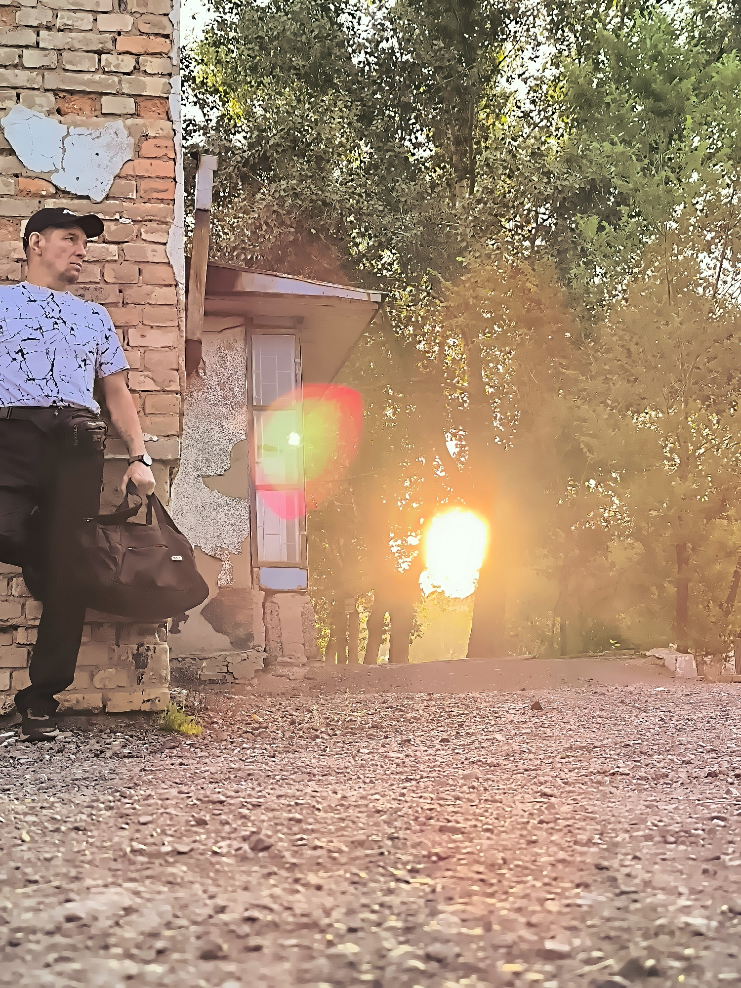 A man wearing a black baseball cap, a white t-shirt with a black abstract pattern, and dark pants stands leaning against a crumbling brick wall. He is holding a dark duffel bag in his right hand. The background features trees with sunlight filtering through the leaves, creating a warm, hazy effect. A window with a blue frame is visible on the wall behind him. The ground is covered in gravel. The overall scene appears to be outdoors, possibly in an urban or semi-urban setting.