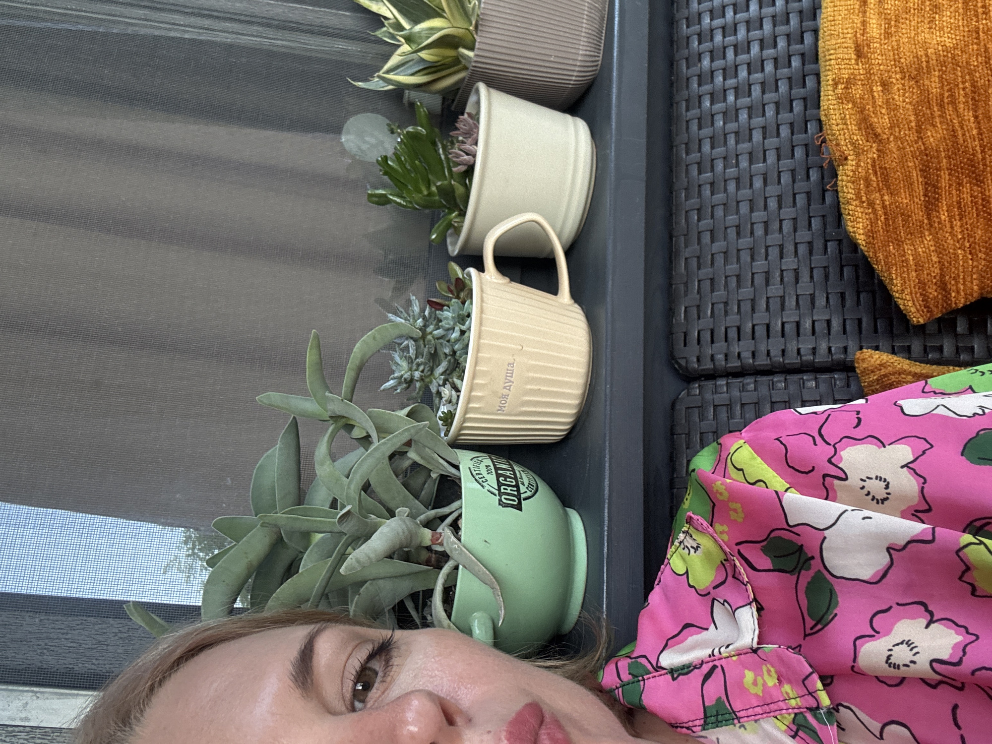 This media file shows a close-up of a person lying down outdoors, with several potted plants visible. The person is lying on a woven patio chair, partially covered by a bright pink floral-patterned garment and an orange textured throw. The plants are in different colored pots: a beige ribbed pot at the top, a cream-colored mug, a light green mug with "CERTIFIED ORGANIC" and other text, and a larger succulent in the foreground. The background appears to be a patio setting, with a textured grey surface and a window screen. The lighting suggests it is daytime, possibly sunny. The person's face is partially visible, with their eyes looking upwards and a neutral or slightly relaxed expression. The overall impression is one of leisure or relaxation in an outdoor setting. The visible text on the green mug is "CERTIFIED ORGANIC". The text on the cream mug appears to be in Cyrillic script. The location is specified as Sochi, Russia.