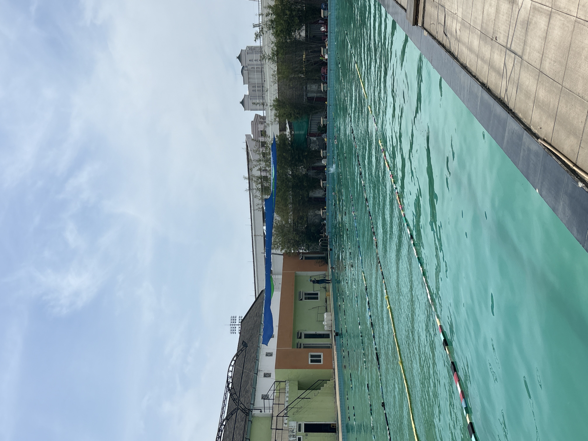 This is an outdoor shot of a swimming pool in Nha Trang Ward, Vietnam, on a cloudy day. The pool, with turquoise water, is divided into lanes by floating ropes with colorful markers. A tiled walkway is visible on the right side of the frame. On the left, a building with green and orange accents houses changing rooms or offices. In the background, trees and modern buildings are visible. Several people are in the pool, swimming or standing near the edge. One person in a blue uniform appears to be cleaning the pool area. The atmosphere is serene, with no indication of any particular event or activity beyond routine use of the pool.