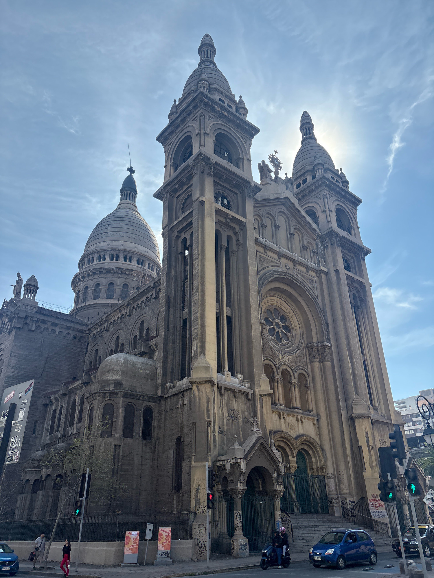 The image captures a low-angle view of the imposing Basilica of the Sacred Heart of Jesus (Basílica del Sagrado Corazón de Jesús) in Santiago, Chile, on a clear day with some wispy clouds. The grand stone structure features multiple towers, spires, and a large central dome, all adorned with intricate architectural details. Sunlight streams from behind the right-hand tower, creating a slight lens flare.
In the foreground, a street scene unfolds. A blue car is parked on the right, while a black taxi is visible behind it. A dark motorcycle with two occupants, one wearing a pink helmet, is in motion on the road. Further left, a blue car is also present. On the sidewalk, two individuals, a woman in red pants and a man in a grey shirt, are walking towards the left.
The building's facade exhibits signs of age, with some graffiti visible on lower sections and fencing. Traffic lights are present, with the green light illuminated. The overall impression is of a historical and prominent landmark within an urban environment.