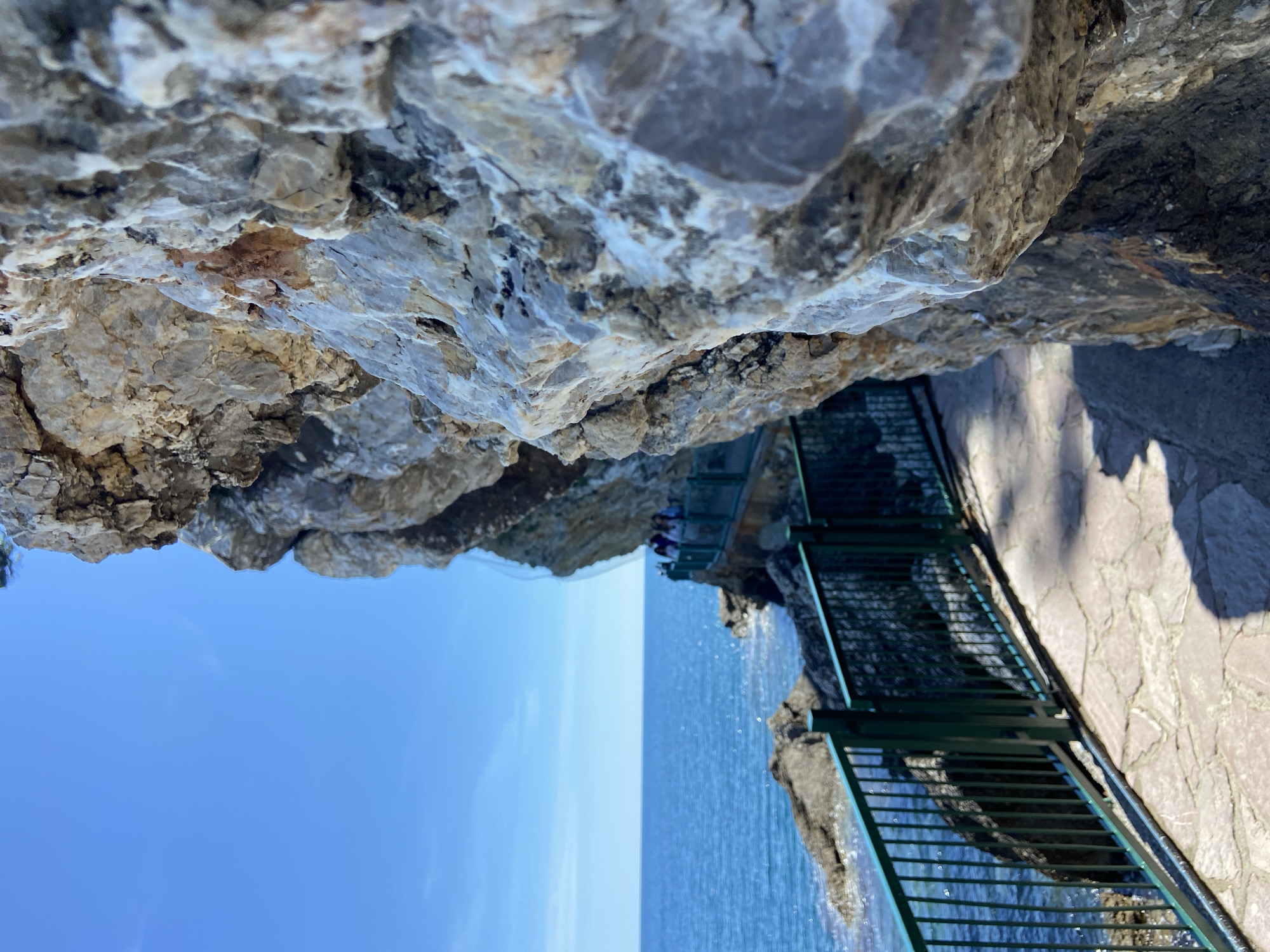 The image depicts a rocky cliffside in Budva, Montenegro, overlooking the Adriatic Sea. The viewpoint is from beneath a rock overhang, looking out to the sea. A walkway with a green metal railing is visible on the right side of the frame. A group of people is gathered on a distant part of the walkway, near the sea. The sky is clear and blue. The sea is calm with blue water. The rocks are light gray in color. The walkway is made of light-colored stone.