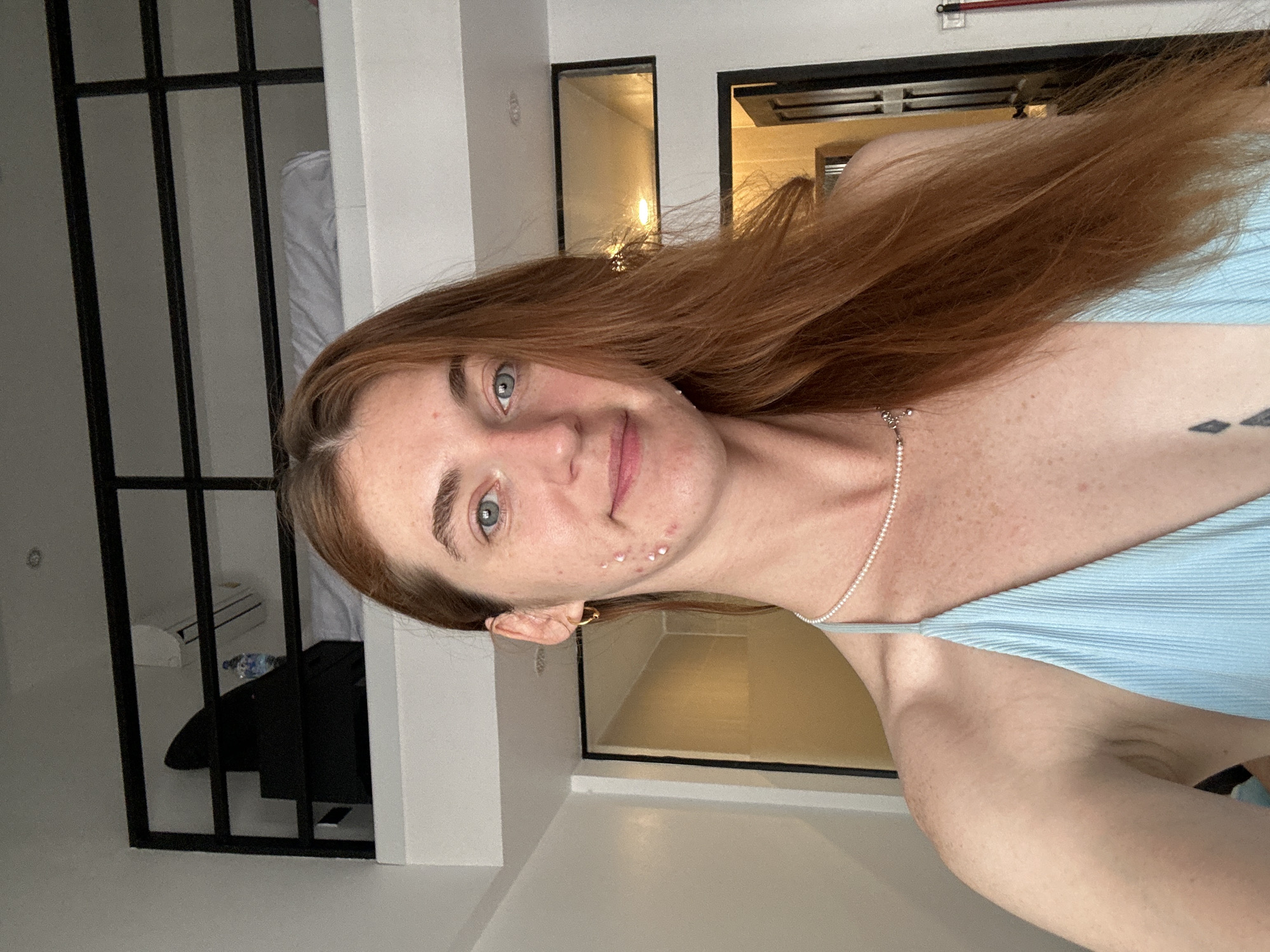A young woman with reddish-brown hair and light blue eyes is featured in a close-up selfie, looking directly at the camera with a subtle smile. She has noticeable freckles across her face and several small white dots on her right cheek. She is wearing a light blue ribbed top and a delicate pearl-like necklace. A dark tattoo is visible on her upper left arm/shoulder.
The image is set indoors, likely in an accommodation in Baan Chaweng, Thailand. In the background to her left, black metal bars suggest a loft bed or room divider, through which a bed with white bedding, a white air conditioner unit, a black piece of clothing, and a water bottle are visible. To her right, white walls and two black-framed windows or mirrors reflect warm indoor lighting, creating an ambient glow. The overall setting appears to be a modern hotel room or apartment. No text is visible in the image.