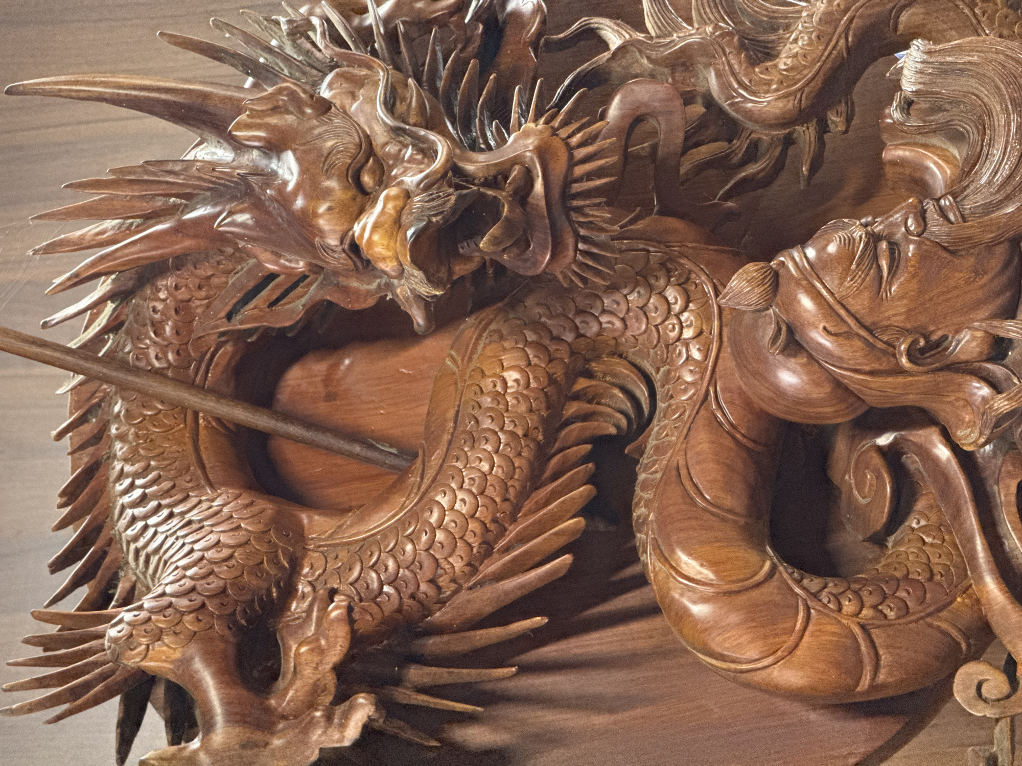This image depicts a detailed wooden carving of a dragon. The dragon is intricately sculpted with scales, sharp claws, and a fierce expression. It appears to be coiled around a human figure, whose face is partially visible. The human has a long beard and is wearing a traditional headdress, suggesting a historical or mythological scene. The carving is made of a reddish-brown wood and is presented against a plain wooden background. The lighting highlights the texture and depth of the carving, emphasizing the craftsmanship. There is no information about the location in the city of Mộc Châu, Vietnam, in the image itself.
