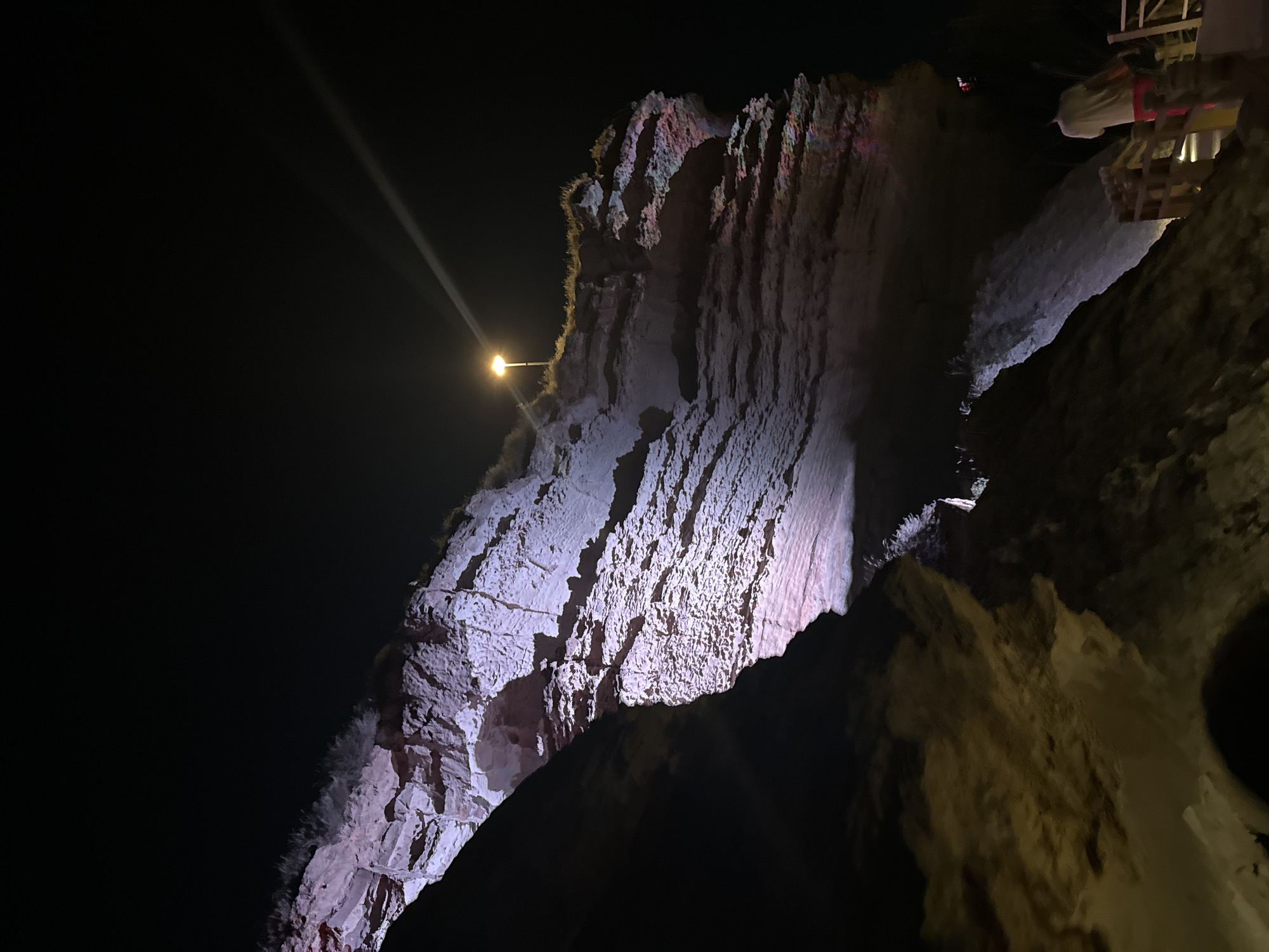 The image depicts a dramatic nighttime scene of the chalk cliffs of Aktau, Kazakhstan, illuminated by artificial lighting. The cliff face is rugged and layered, with visible textures and natural formations. A single street lamp is perched on the edge of the cliff, casting a bright beam of light that creates a strong highlight and a visible light streak across the dark sky. The lower portion of the cliff and the surrounding area are cast in shadow, with some areas showing hints of pink and purple lighting. A person is visible in the lower right corner, standing on a wooden walkway or staircase, facing away from the camera. The overall atmosphere is mysterious and somewhat surreal, highlighting the natural beauty of the cliffs under artificial illumination at night.