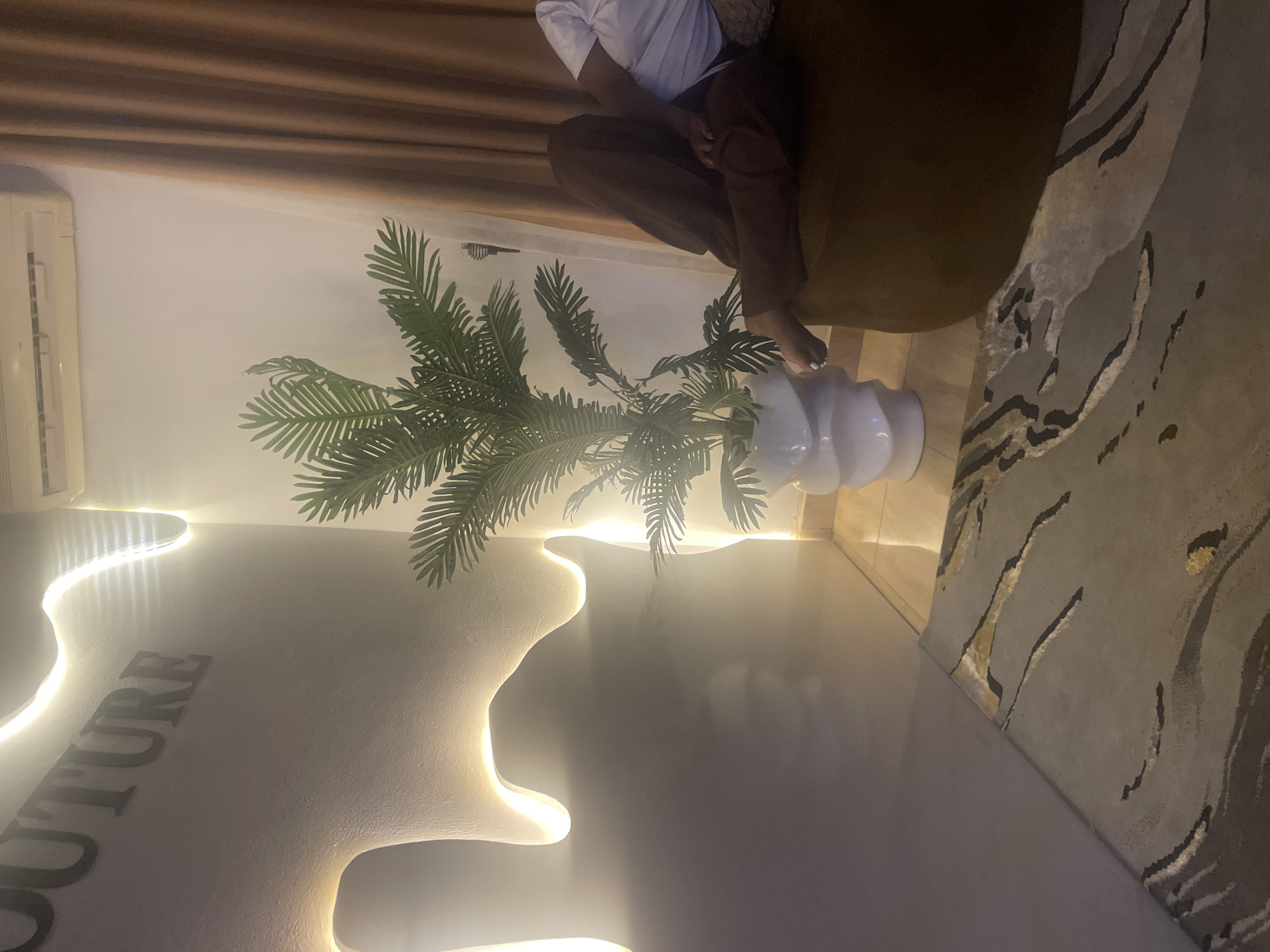 The image captures an indoor scene, likely a modern living room or lounge area, during the evening or night, as indicated by the prominent use of artificial lighting. The location is Dushepe, Nigeria.
A person is visible, seated on a dark brown or olive-green sofa or plush chair, facing away from the camera. Only their lower torso and legs are visible; they are wearing a white t-shirt and dark brown trousers. Their bare feet are relaxed on the floor, and their toenails have white polish.
The room features a contemporary design. A large, artificial palm-like plant with vibrant green fronds stands in a stylish white, multi-tiered vase on the floor, positioned near the seated person. The walls are a light, neutral color, possibly white or off-white. Unique, organic-shaped wall panels are a dominant feature, integrated with hidden LED strip lighting that casts a warm, soft glow, highlighting their contours. Part of a word is visible on one of these illuminated panels, spelling "OUTURE," suggesting "COUTURE."
An air conditioning unit is mounted on the wall in the upper left section of the image. Dark brown curtains or blinds are drawn across the upper part of the wall, possibly covering a window. The floor is partially covered by a modern rug featuring abstract, curvilinear patterns in shades of grey, black, and hints of light brown or gold. The overall ambiance is cozy and sophisticated, emphasized by the indirect lighting and natural elements like the plant.