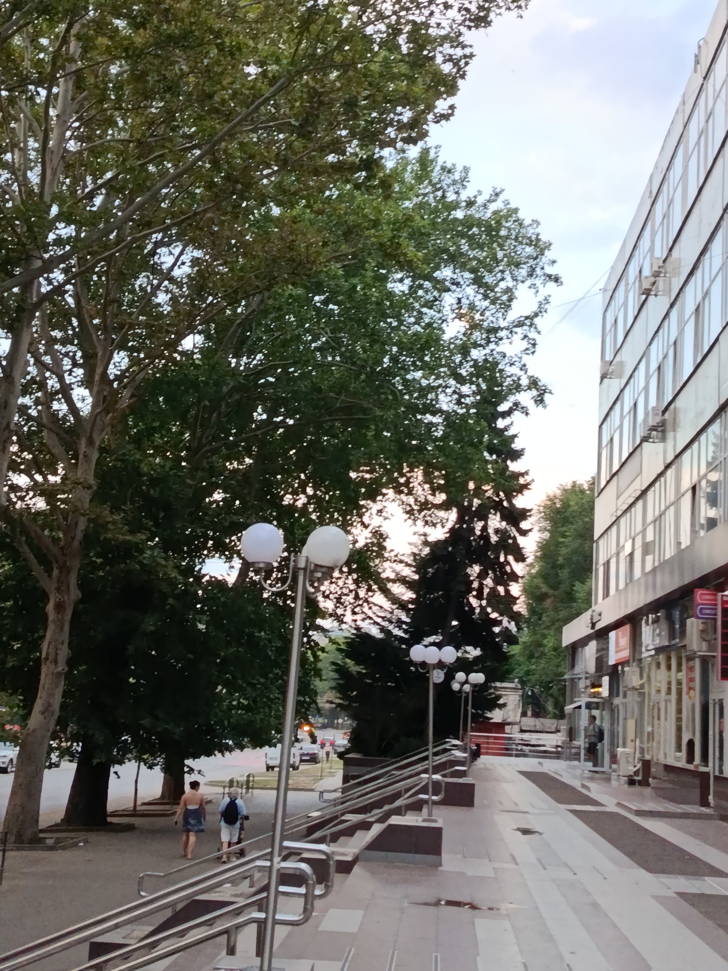 The image captures a street scene in Chișinău, Moldova, on a cloudy day. On the right side of the frame, a multi-story modern building with reflective glass windows dominates the view. Air conditioning units are visible mounted on the exterior walls. Below the windows, storefronts with various signs and entrances are present. A wide, paved walkway with steps and metal railings leads away from the foreground towards the building. To the left, a large tree with lush green foliage casts shadows on the sidewalk and a paved street. Several cars are parked along the street. Two individuals are walking away from the camera on the sidewalk, one wearing a dark patterned dress and the other a light blue shirt and white shorts, carrying a backpack. Further down the walkway, a few more people are visible near the building entrances. Streetlights with multiple white globes are positioned along the walkway. In the background, more trees and some architectural elements can be seen.