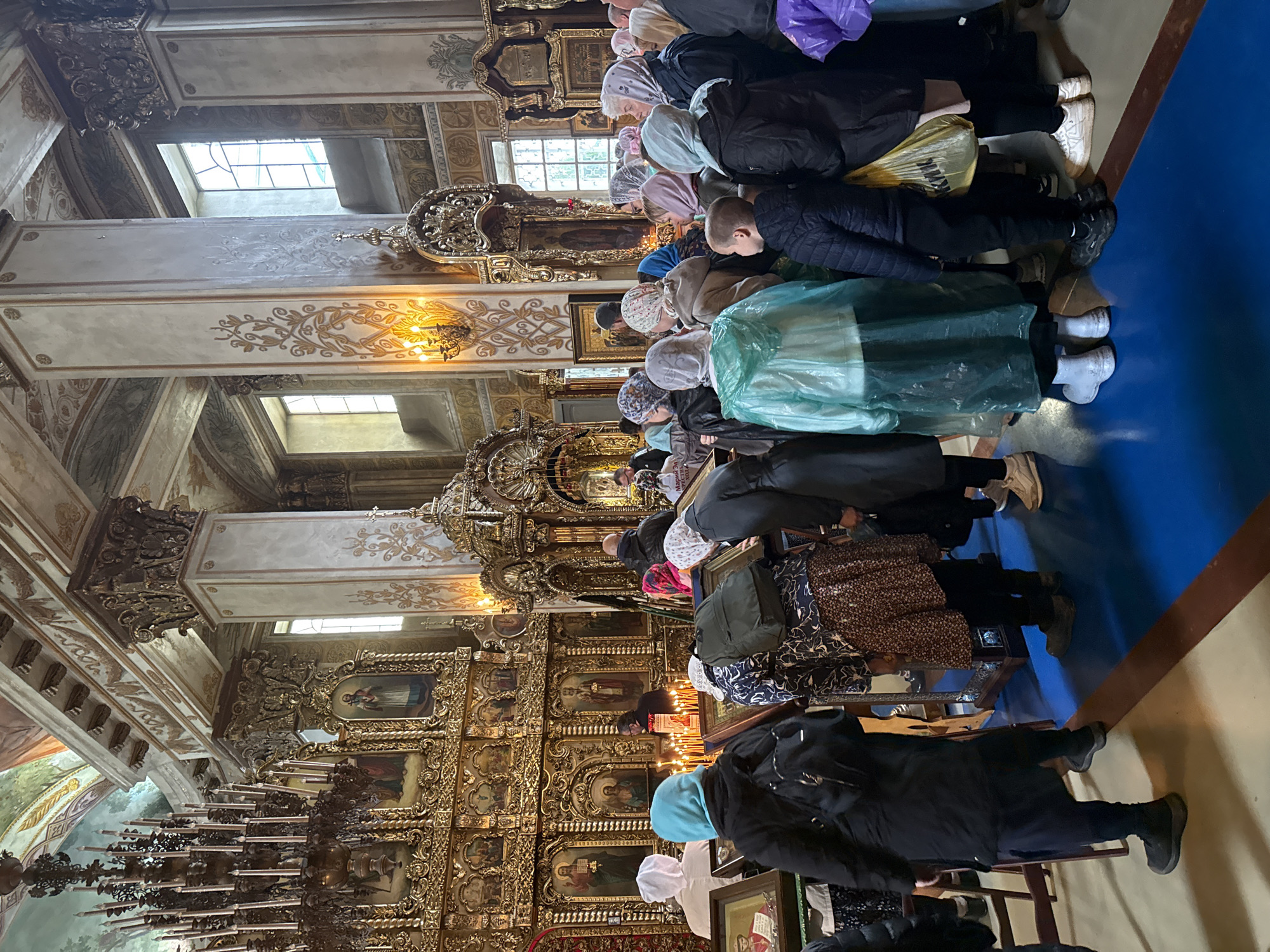 The image depicts the interior of an ornate Orthodox church, likely within the city of Раифа, Russia. A group of people, predominantly women, are gathered in front of a large, gilded iconostasis adorned with multiple religious icons and lit candles. Many are wearing headscarves, and some are kneeling or bowing, engaging in prayer or veneration. The church architecture is richly decorated with columns featuring intricate carvings and gold accents, and the ceiling is painted with frescoes. Sunlight streams in through tall, arched windows. The floor is covered in a vibrant blue carpet. The overall atmosphere suggests a religious service or a period of devotion.