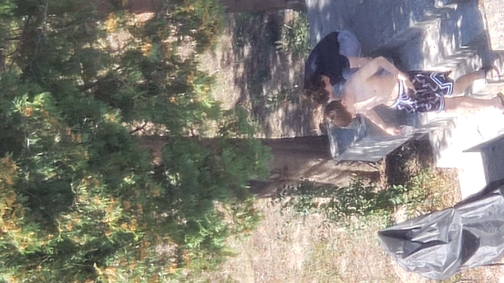 The image depicts two individuals outdoors, beneath the canopy of a large tree with green leaves and small yellow flowers. The ground appears to be dry and covered in sparse vegetation. In the foreground, a large black plastic bag is visible. To the right, two young males are on a concrete structure that resembles steps or a tiered seating area. One individual, shirtless and wearing patterned shorts, is holding a clear cup and appears to be pouring liquid. The second individual, wearing a dark t-shirt and jeans, is crouched down. The setting suggests a warm, outdoor environment. The location within Prešov, Slovakia, is not discernible from the image itself.