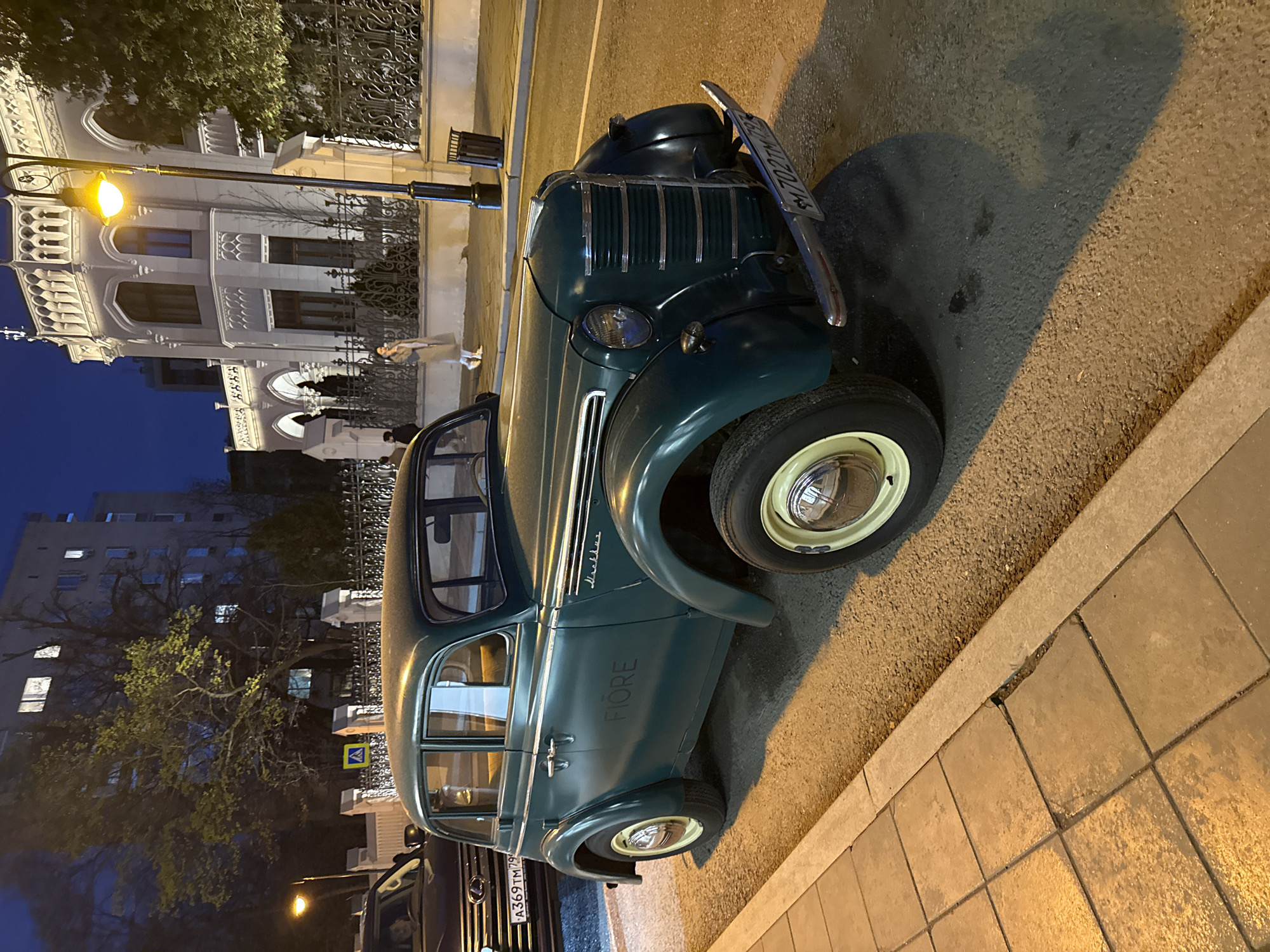 At night, a green vintage car with a white-walled tire is parked on the street in Moscow, Russia. It appears to be a Moskvich 400. The license plate on the car is A369TM 799. In the background, there is a building with a white façade, and a street lamp is visible near a wrought-iron fence. A person is walking near the building. Another car is parked behind the vintage vehicle.