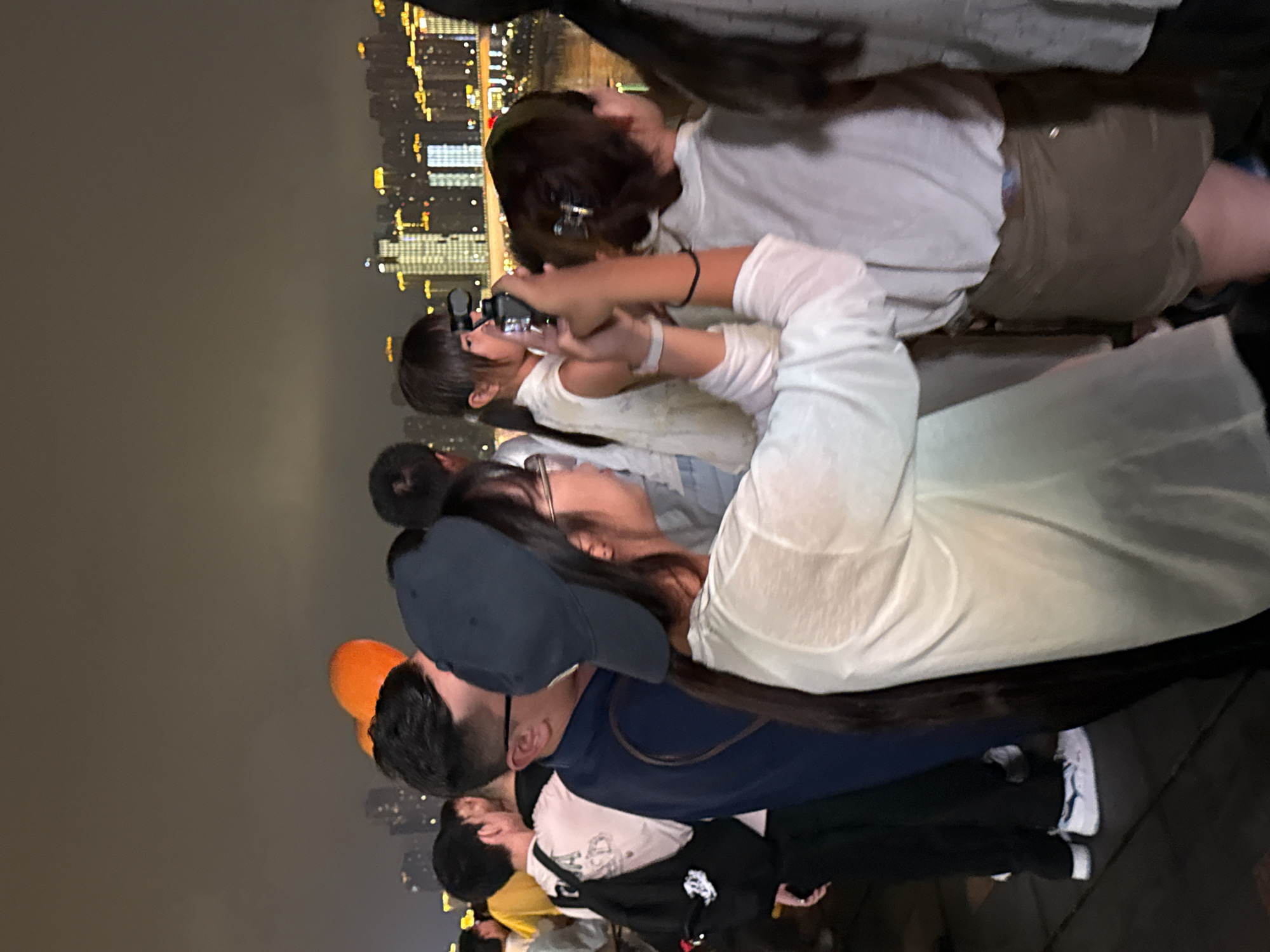 The image depicts a nighttime scene with a crowd of people gathered on a waterfront promenade in Yuzhong District, China, likely near the Jialing River. The background features a brightly lit cityscape with numerous tall buildings. In the foreground, several individuals are visible, some looking out towards the city lights, while others are engaged with mobile devices. A young girl is holding up a phone to take a picture or video, with a woman beside her also looking towards the city. Another woman in the foreground is wearing a blue baseball cap and a white long-sleeved shirt, and appears to be looking to the side. The ground appears to be a dark, paved surface. The overall atmosphere suggests a public gathering or tourist attraction.