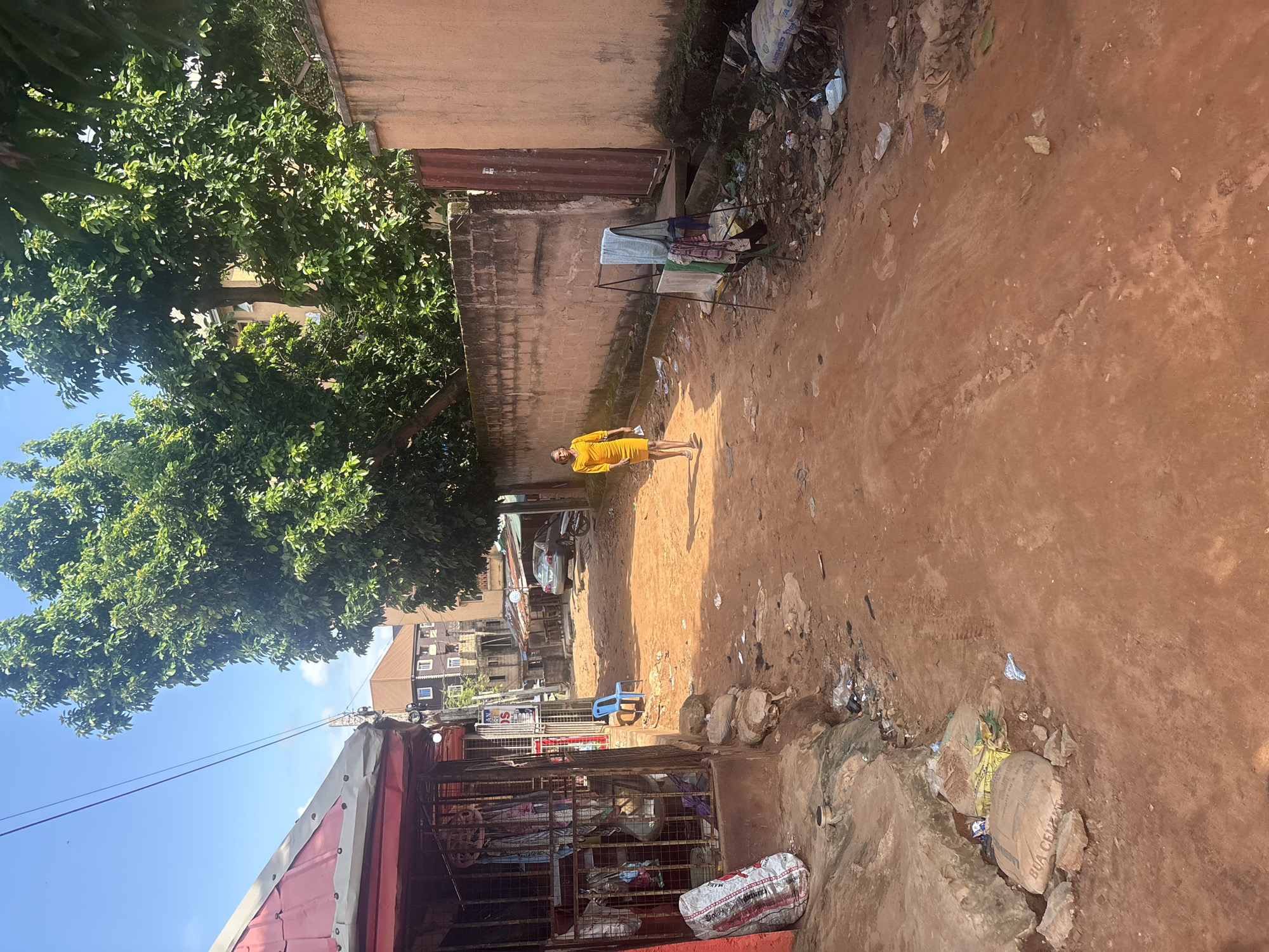 In Nkpor, Nigeria, a woman walks down a dirt road lined with buildings and lush green trees. The road is uneven and appears to be a thoroughfare for local commerce. On the left, a small shop with a red roof and metal bars displays various items, including clothing and what looks like bags of cement. A blue plastic chair sits outside the shop. Further down the road, a light-colored car and a motorcycle are parked. On the right side of the road, a tall, textured wall stretches along the scene, with a metal gate visible. A clothes drying rack stands in front of the wall, holding several garments. The ground is littered with debris, including plastic bags and discarded items, and a small, open drain runs along the base of the left-hand buildings. The sky is bright blue with scattered clouds, indicating a sunny day. The overall impression is of a vibrant but somewhat unkempt urban environment.