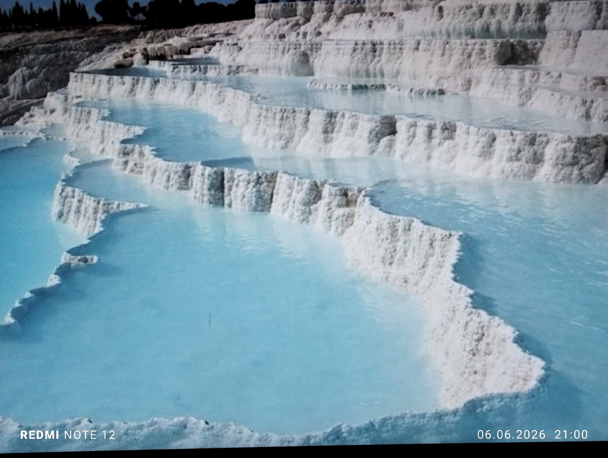 The image displays an expansive outdoor scene featuring tiered natural formations located in the city of Burgas, Bulgaria. White, calcified terraces, characterized by their irregular shapes and rough textures, descend in a series of steps. Each step forms a basin filled with clear, tranquil, light blue water. The water pools vary in size and shape, creating a visually striking contrast with the bright white mineral deposits. In the upper background, a dark, tree-lined ridge is visible against a deep blue sky. No individuals or animals are present within the frame. The lower left corner of the image displays a "REDMI NOTE 12" watermark, while the lower right corner shows a timestamp of "06.06.2026 21:00".