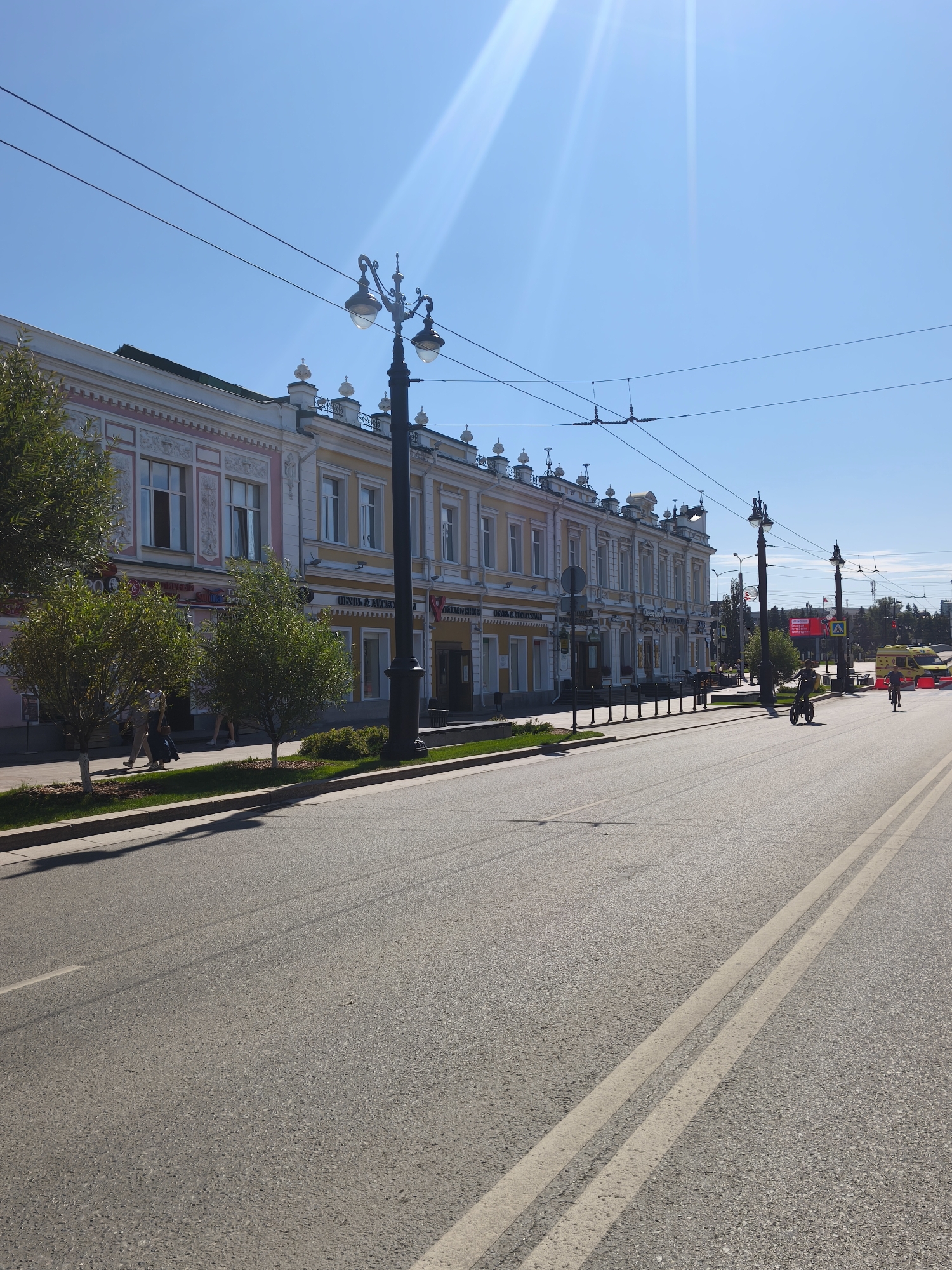 The image depicts a street scene in Omsk, Russia, likely during daytime given the bright sunlight and clear blue sky. A multi-story building with ornate architecture, painted in pale yellow and pink, lines the left side of the street. Several trees with green foliage are planted along the sidewalk in front of the building. The street itself is paved and appears to be a multi-lane road with painted white lines.
Several individuals are visible. On the sidewalk, two people are walking, one wearing a patterned top and dark skirt, the other in light-colored trousers. Further down the sidewalk, a few more people are walking, some appearing to be in groups. On the street, a person is riding a bicycle or electric scooter, and another person is cycling on a bicycle. An ambulance, with yellow and red markings, is parked further down the street. Street lamps and overhead wires are also present. The overall atmosphere suggests a typical urban environment.