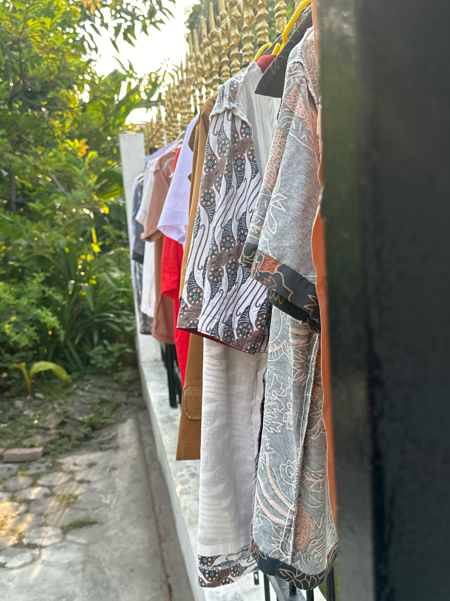 The image captures a close-up view of several batik shirts hanging on a clothesline, likely outdoors in a residential area. The shirts exhibit traditional batik patterns in various colors and styles, with the foreground showing the intricate designs of the fabric. In the background, lush green foliage suggests a tropical or subtropical environment. The sunlight filtering through the leaves indicates it is either morning or late afternoon. The setting appears to be a secured area, possibly a yard or balcony, with a decorative fence and a concrete ledge. While the specific location is not visible, the presence of batik clothing and the tropical greenery suggests a setting within Indonesia, and potentially within the city of Mojokerto, known for its cultural heritage and warm climate. There are no people or animals visible in the frame.