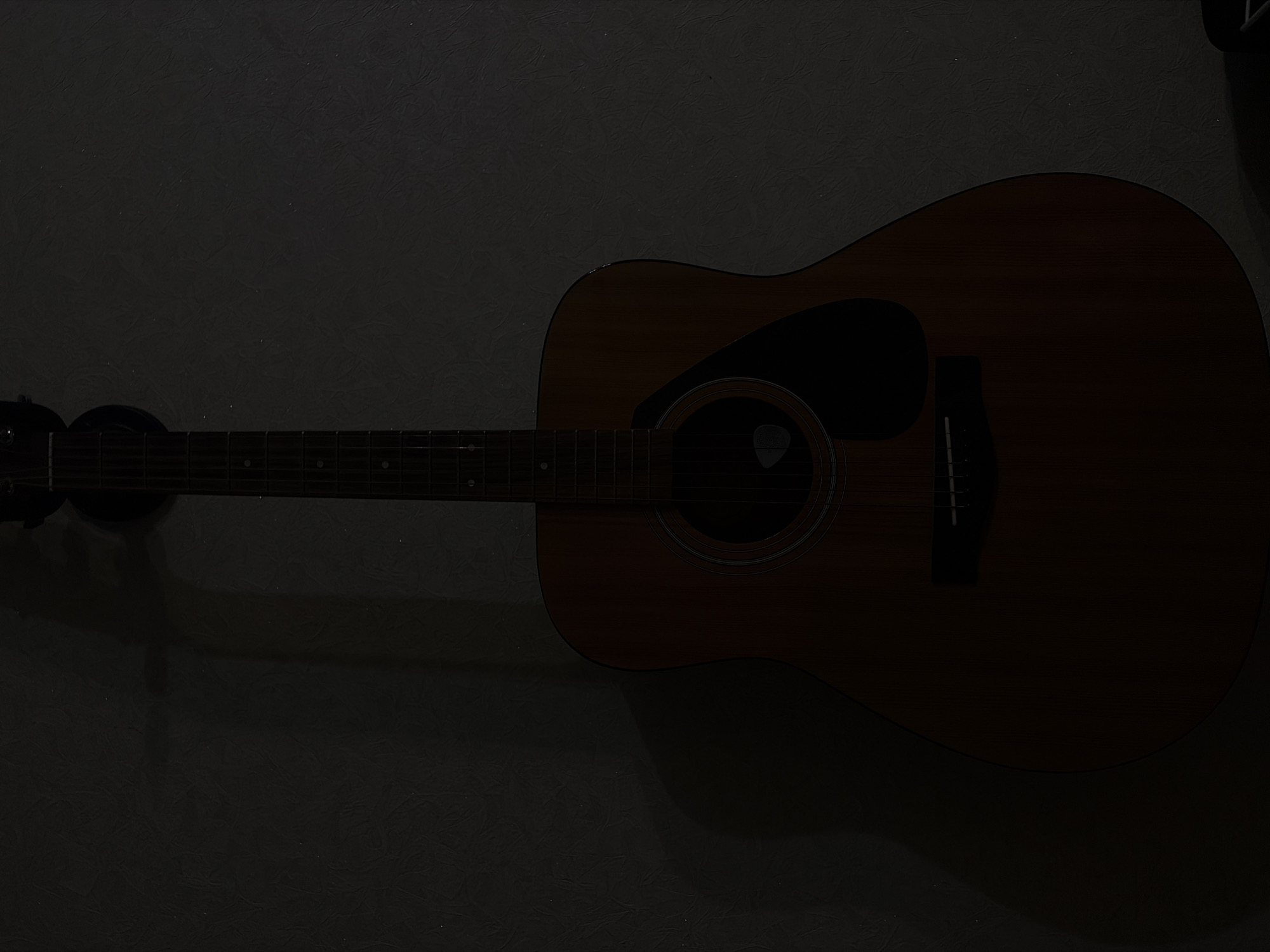 This interior scene, located in Tuymazy, Russia, displays a full-body view of an acoustic guitar hanging vertically against a textured, light-colored wall. The guitar features a reddish-brown wooden body with a black, teardrop-shaped pickguard positioned below its circular soundhole. A dark-colored guitar pick is lodged partially within the soundhole. The fretboard and neck are dark, with several visible fret markers. The headstock is largely obscured by shadow at the top of the frame. The scene is dimly lit, and a prominent shadow of the guitar is cast to its left on the wall, indicating a light source from the right. The wall exhibits a subtle, irregular textured pattern. A dark, indistinct object is partially visible in the lower right corner of the frame.