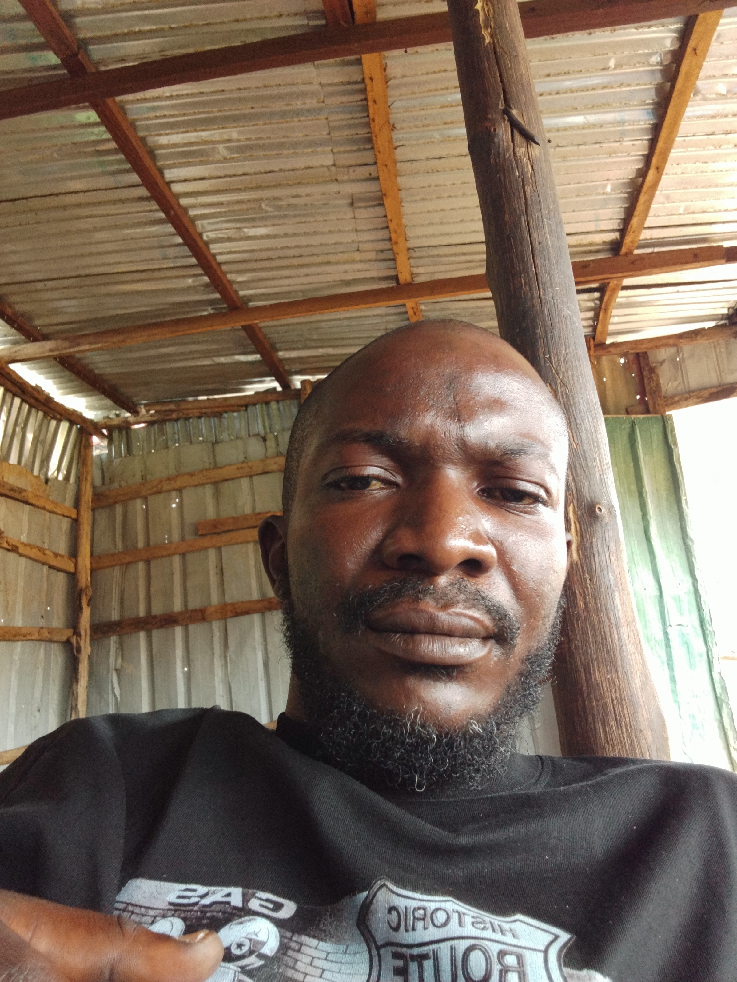 A close-up, front-facing selfie of a Black man with a shaved head and a beard is captured from the chest up. He is wearing a black t-shirt with a graphic design that appears to be a shield-shaped emblem with text, possibly a route marker. The man is leaning against a rough-hewn, vertical wooden post, which is a prominent element in the scene.
Behind him, the setting is an open-air structure with a corrugated metal roof supported by wooden beams. The walls appear to be made of corrugated metal and some wooden planks, suggesting a rustic or unfinished construction. The lighting indicates it is daytime, possibly bright, with sunlight filtering in, casting some shadows and highlighting the textures of the materials.
The man's expression is neutral, and his gaze is directed towards the camera. The overall impression is informal, perhaps taken in a rural or outdoor workspace. The visible text on his shirt reads "ROUTE" and "HISTORIC" in a stylized font, with some other illegible text. The image has a candid feel, as if captured spontaneously. The context of Durimi, Nigeria, suggests this is likely an outdoor setting within a community there.