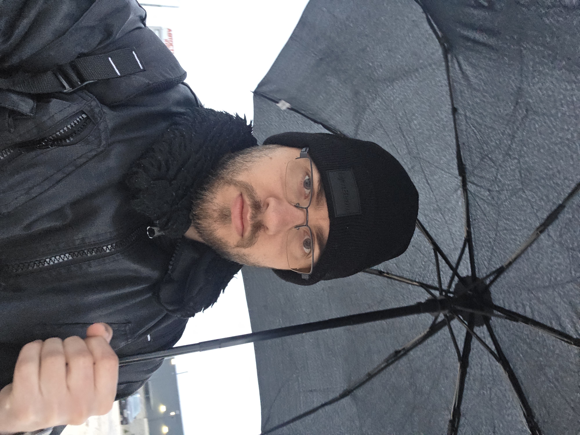 A person is holding a black umbrella. The person is wearing a black jacket and a black hat. The person also wears glasses. The photograph may have been taken in Plovdiv, Bulgaria.