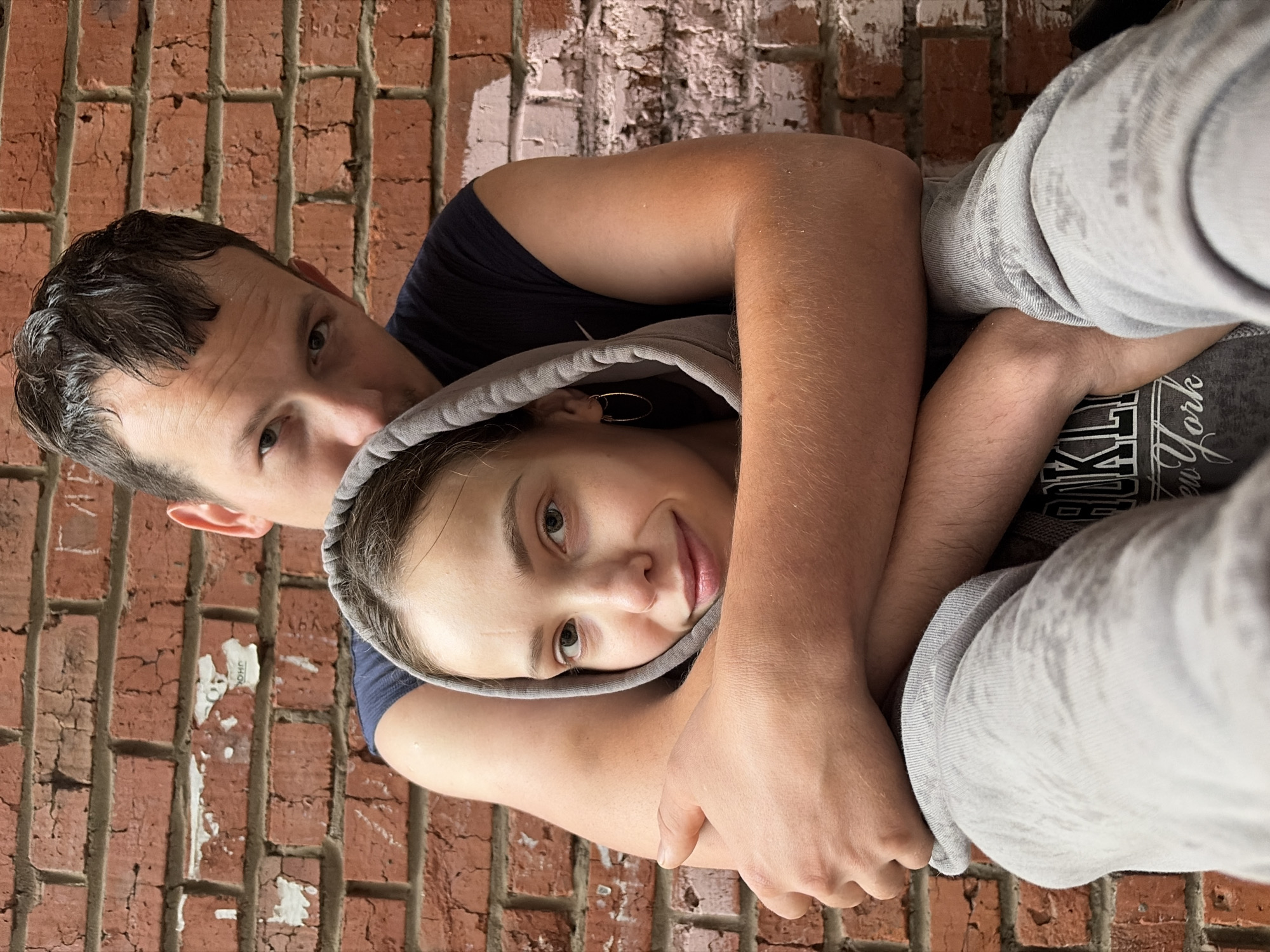 A man and a woman are embracing in front of a brick wall. The man is behind the woman, with his arms wrapped around her. Both are wearing casual clothing. The woman has a hood pulled up over her head and is looking towards the camera. The man is also looking towards the camera, with a slight smile. The brick wall appears to be weathered and worn, with some peeling paint. The image does not provide any specific details about the city of Noviy, Russia.