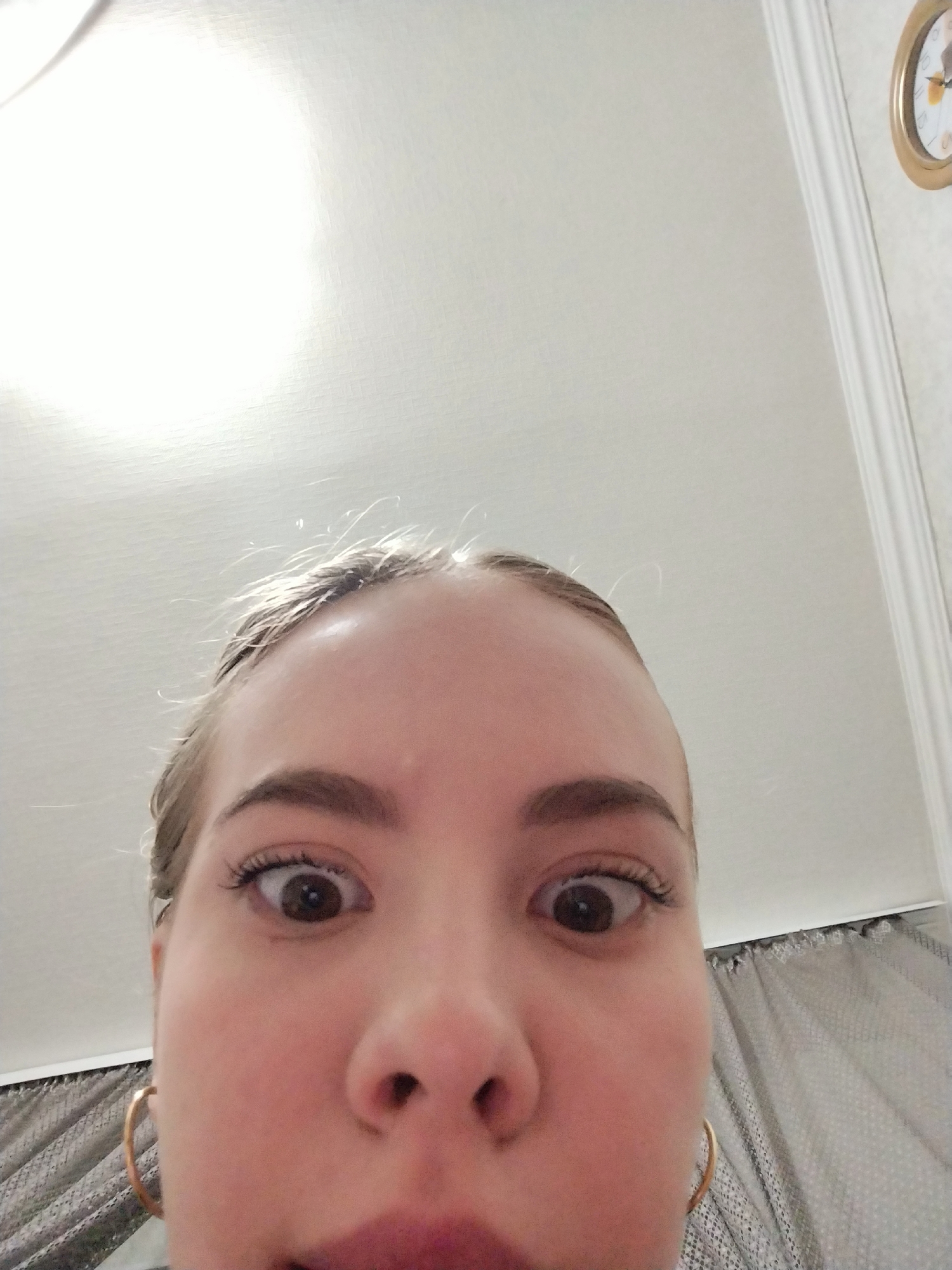 In a close-up, low-angle selfie, a young woman with light brown hair pulled back is looking directly into the camera. Her eyes are wide open, and her mouth is slightly agape, suggesting surprise or a playful expression. She is wearing large hoop earrings. The background is a plain white ceiling with a subtle texture and a faint light source, indicating an indoor setting. To the right, a portion of a wall with decorative molding and a round clock with a gold frame is visible. The overall lighting is bright, casting a soft glow on the subject's face.