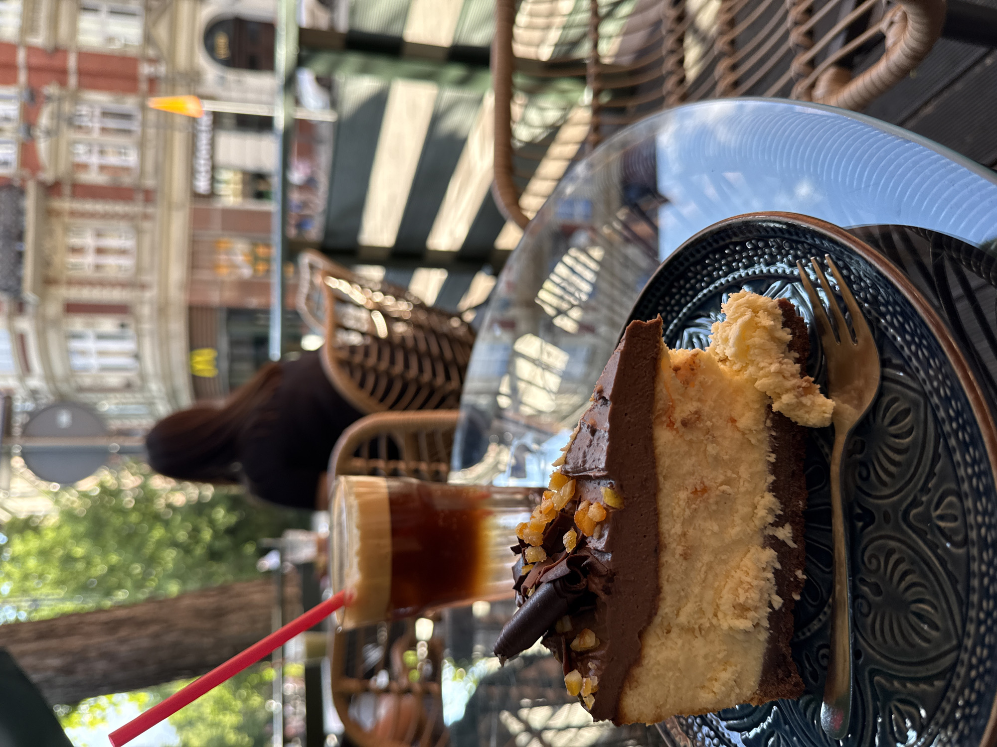 The image depicts an outdoor café scene on a bright day, focused on a dessert and drink in the foreground with an urban background.
A slice of cheesecake with a dark chocolate crust, a light-colored dense cake layer, and a dark chocolate ganache topping occupies the center-left. It is adorned with dark chocolate curls and scattered orange candied zest. A portion has been consumed from the front, revealing the interior texture. The cake rests on a dark blue/green plate with an intricate embossed pattern. A gold-toned fork is placed on the plate beside the cake.
To the right and slightly behind the cake, a clear glass contains a layered iced coffee with a dark brown base liquid and a lighter brown foam head. A red plastic straw extends from the drink.
These items are situated on a clear, reflective glass-topped table, revealing reflections of the sky and surroundings.
In the midground, out of focus, a person with dark hair is seated facing away from the camera, wearing a dark top, at another rattan-style café chair and table. Multiple wicker/rattan chairs are visible around the tables.
The background shows a blurred European-style street scene. A multi-story building with red brick walls, white ornate window frames, and several windows is prominent. A partially visible McDonald's golden arches logo is discernable on the ground floor of this building. To the right, a faint yellow/orange flag-like object is visible. Lush green foliage from trees lines the street, and a dark tree trunk is visible on the left. A dark-colored slatted railing separates the cafe area from the street. The precise city and country remain unknown, but the architecture suggests a European urban setting.