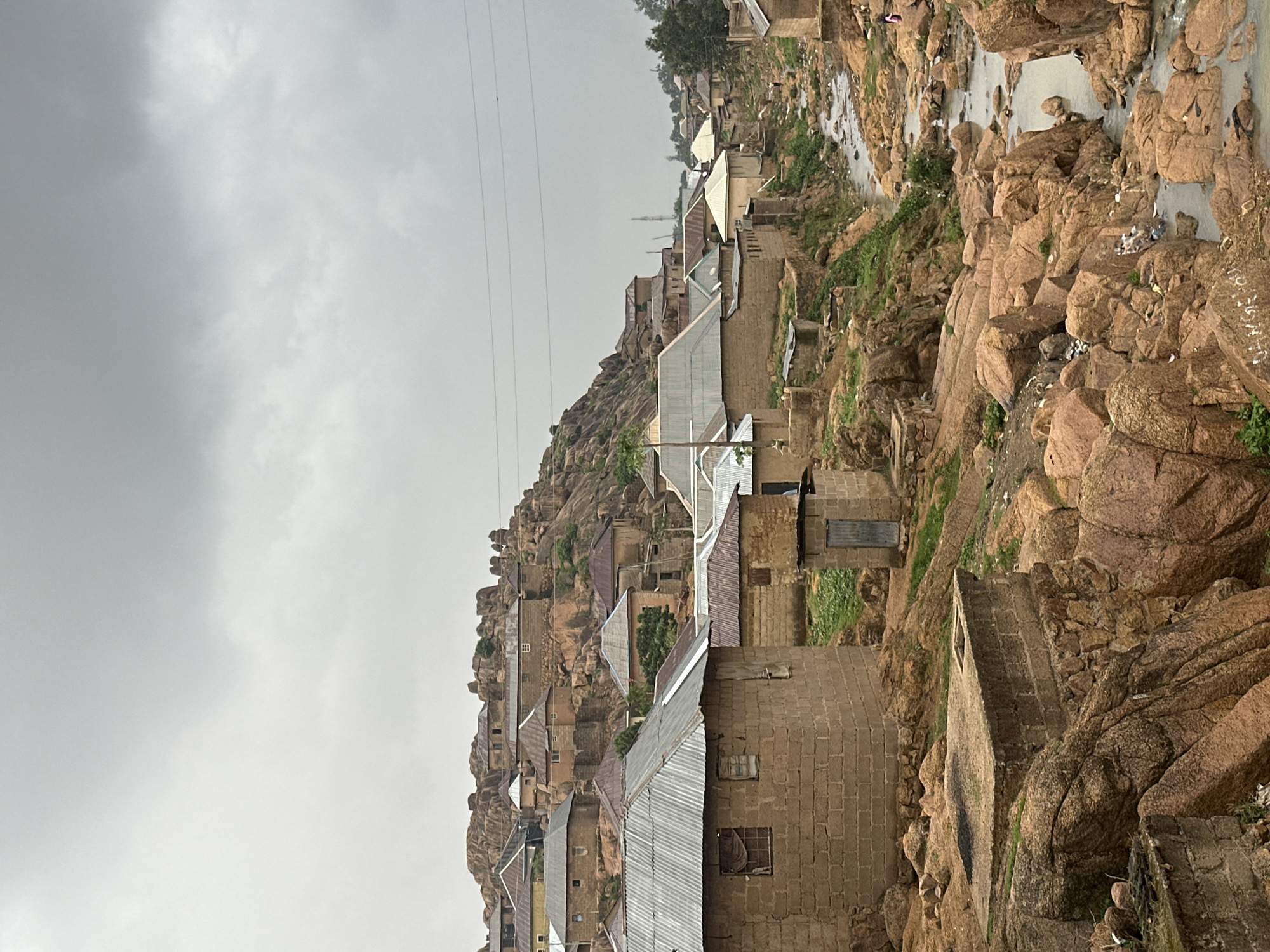The image depicts a settlement nestled among large, reddish-brown rock formations under an overcast sky. The scene is dominated by a rugged, rocky landscape, with numerous boulders and outcrops. Houses, constructed primarily from concrete blocks with corrugated metal roofs, are scattered across the rocky terrain, some appearing to be built directly into or against the rock faces.
A narrow river or stream winds through the right side of the frame, flowing between the rocks. The water appears murky, and some debris or litter is visible in and along its banks. Sparse green vegetation grows in patches between the rocks and around the houses. Overhead, several parallel power lines traverse the sky. The overall lighting suggests a cloudy or overcast day, with no direct sunlight. No people or specific human activities are visible. On one of the large rocks near the river on the far right, there are some faint white markings or text, though it is illegible. The architectural style and rocky environment are consistent with urban or semi-urban areas in Jos, Nigeria.