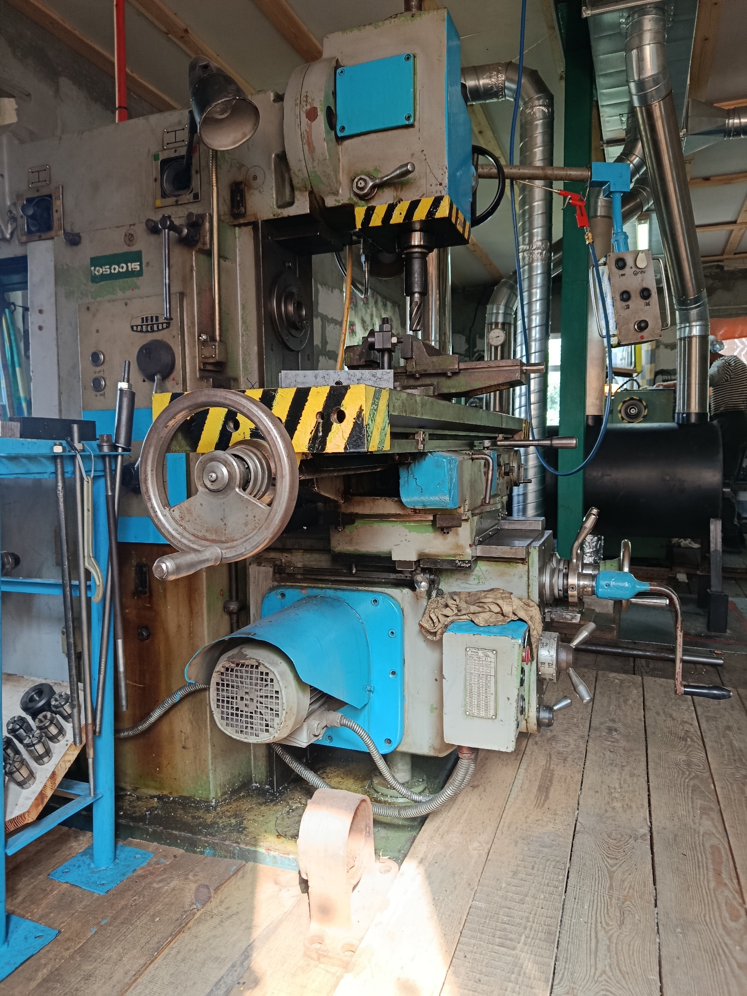 A close-up, eye-level shot captures a large industrial milling machine, predominantly painted in a faded gray and accented with bright blue and yellow-black warning stripes. The machine occupies the central and left portions of the frame, its complex mechanical elements filling the space. A large handwheel with a silver handle is prominently displayed on the machine's side. To the left, a blue metal rack holds various cylindrical metal tools. The machine is positioned on a wooden floor made of wide, weathered planks. In the background, a person wearing a striped shirt can be seen working in the dimly lit industrial space, which features exposed pipes and ventilation systems. The overall environment suggests an operational workshop. The image's details are consistent with a scene in Motyzhyn, Ukraine.