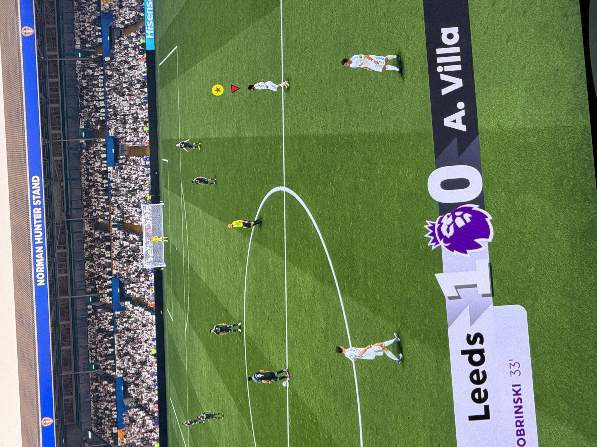 A television screen displays a soccer match between Leeds and Aston Villa (A. Villa). The score is 1-0 in favor of Leeds, with Dobrinski scoring at 33'. The scene is a soccer field with players and the game in progress. Spectators fill the stadium stands. A sign in the background reads "Norman Hunter Stand". The match is not in Burgas, Bulgaria.